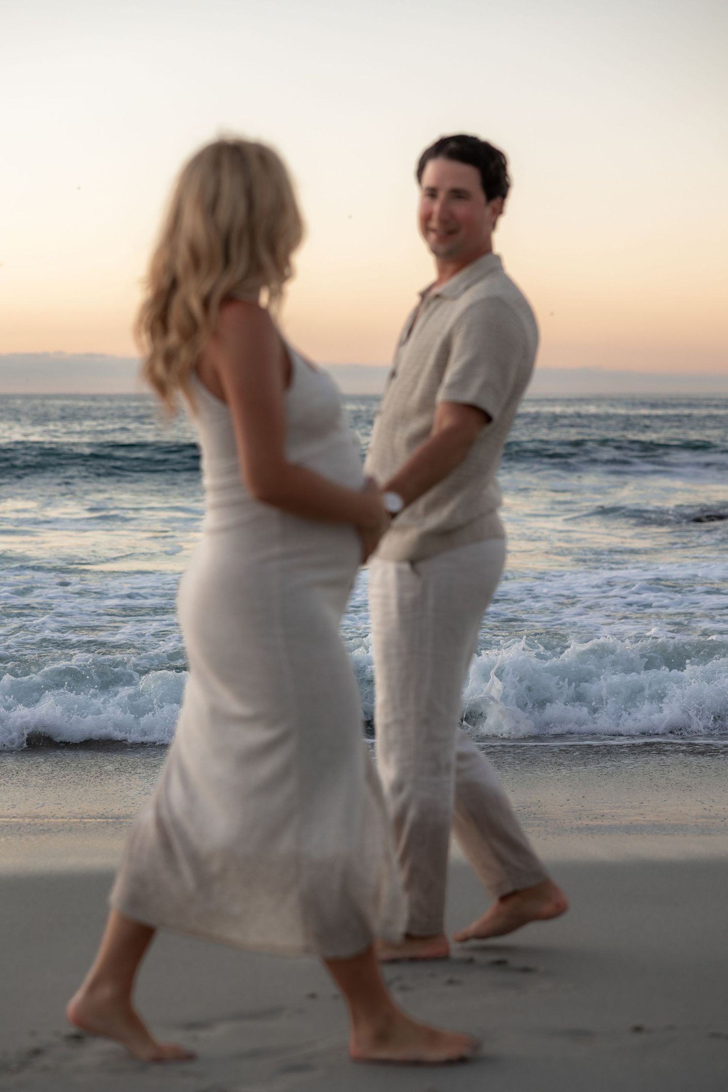 A couple in white outfits dances together on a beach at sunset as waves crash behind them.