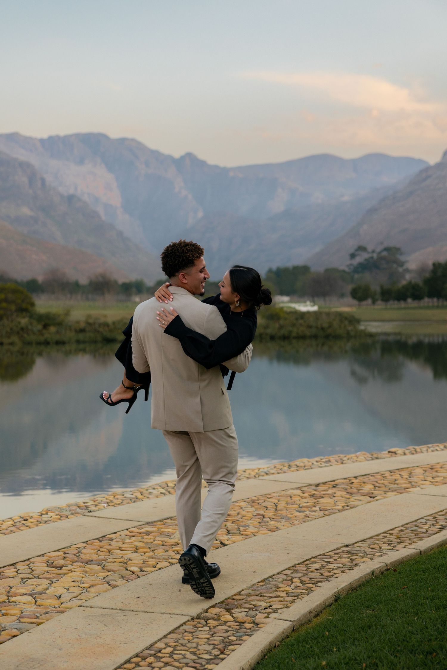 Couple embracing by a scenic mountain lake at dusk, man lifting woman in elegant attire on a stone pathway.