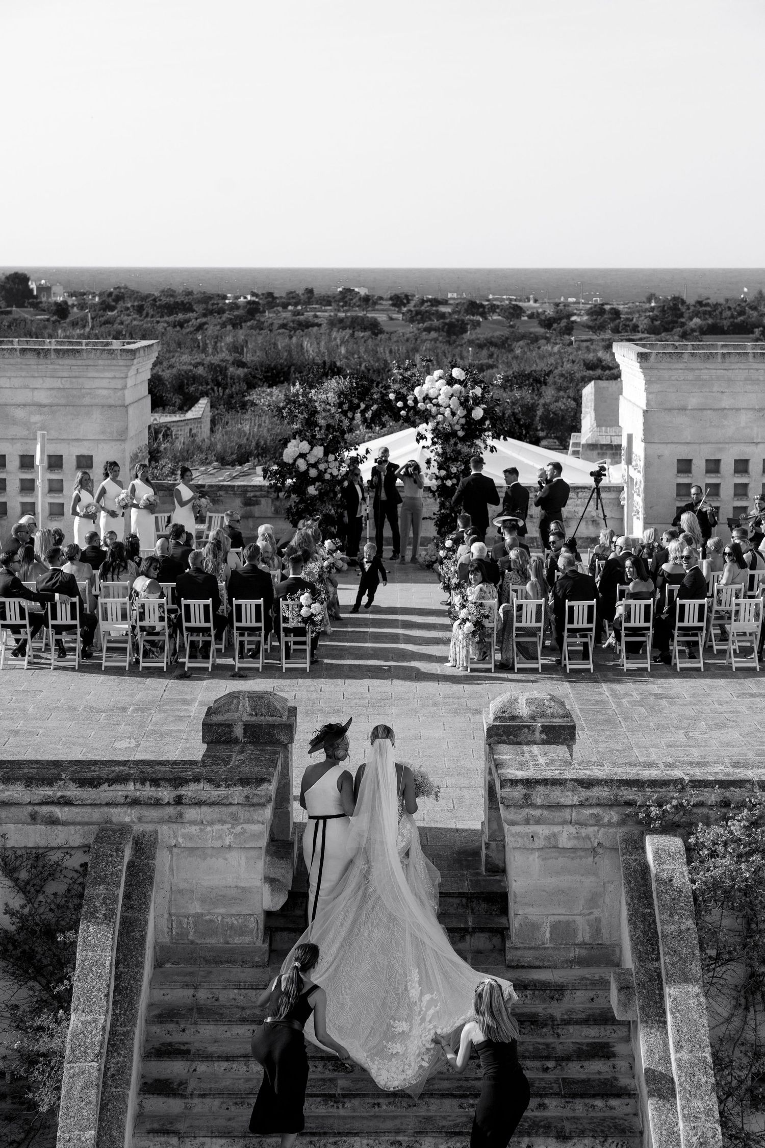 Black and white photo of outdoor wedding ceremony with guests seated on stone steps.