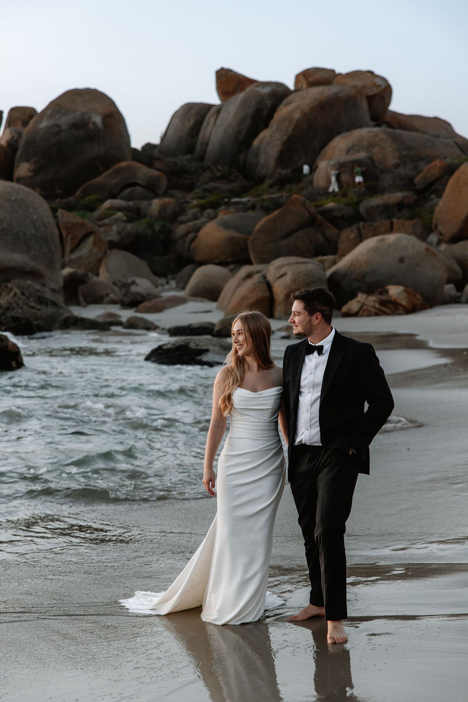 Wedding couple walking along rocky beach shoreline at sunset.