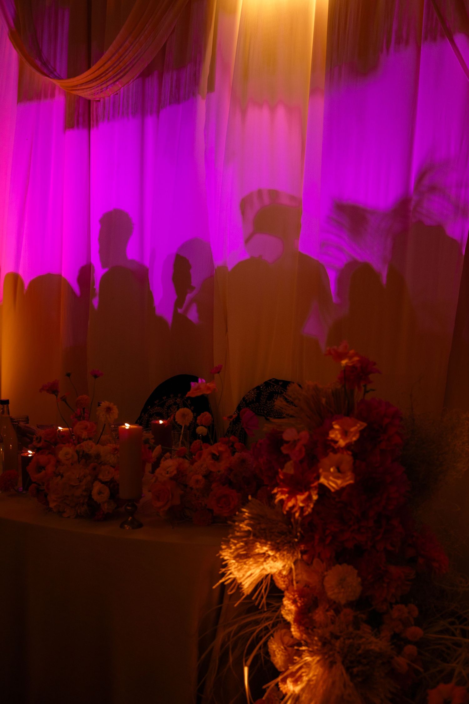 Purple and orange uplighting creates dramatic ambiance at an indoor wedding reception with floral silhouettes.