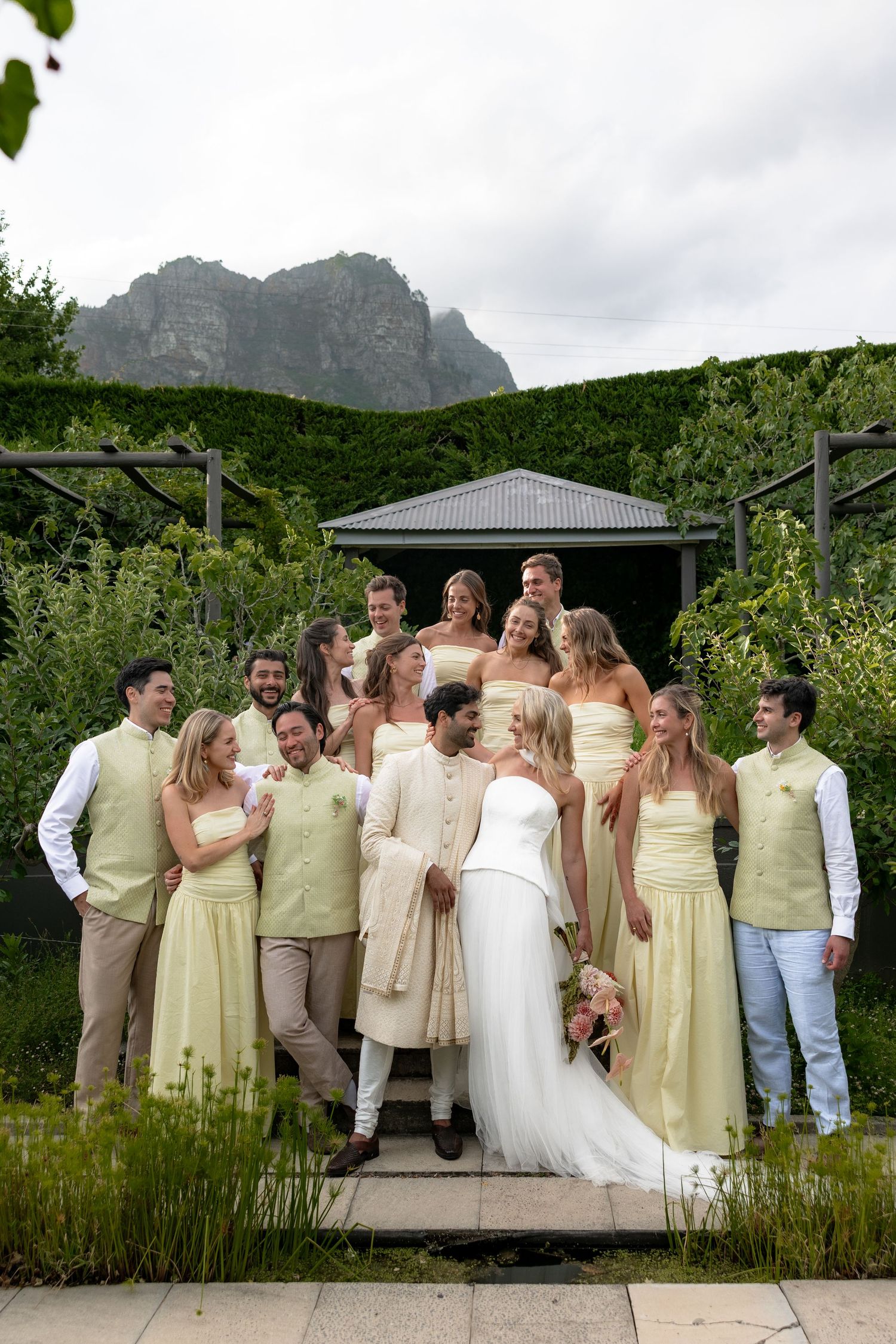 Wedding party in yellow dresses and green vests poses with bride and groom in lush garden with mountains.