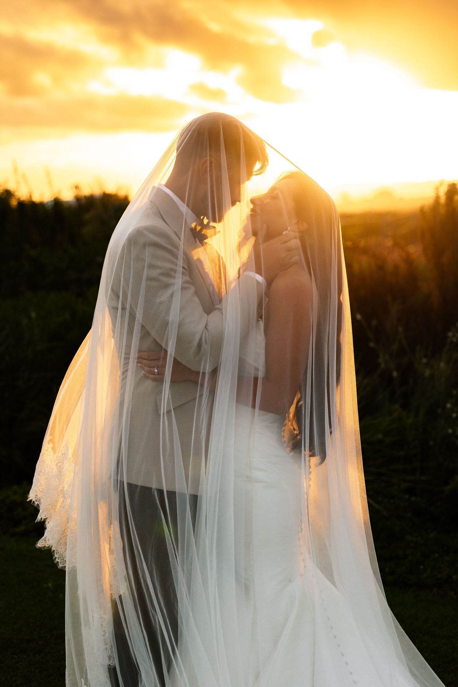 Bride and groom share romantic moment under wedding veil at golden sunset, silhouetted against vibrant orange sky.
