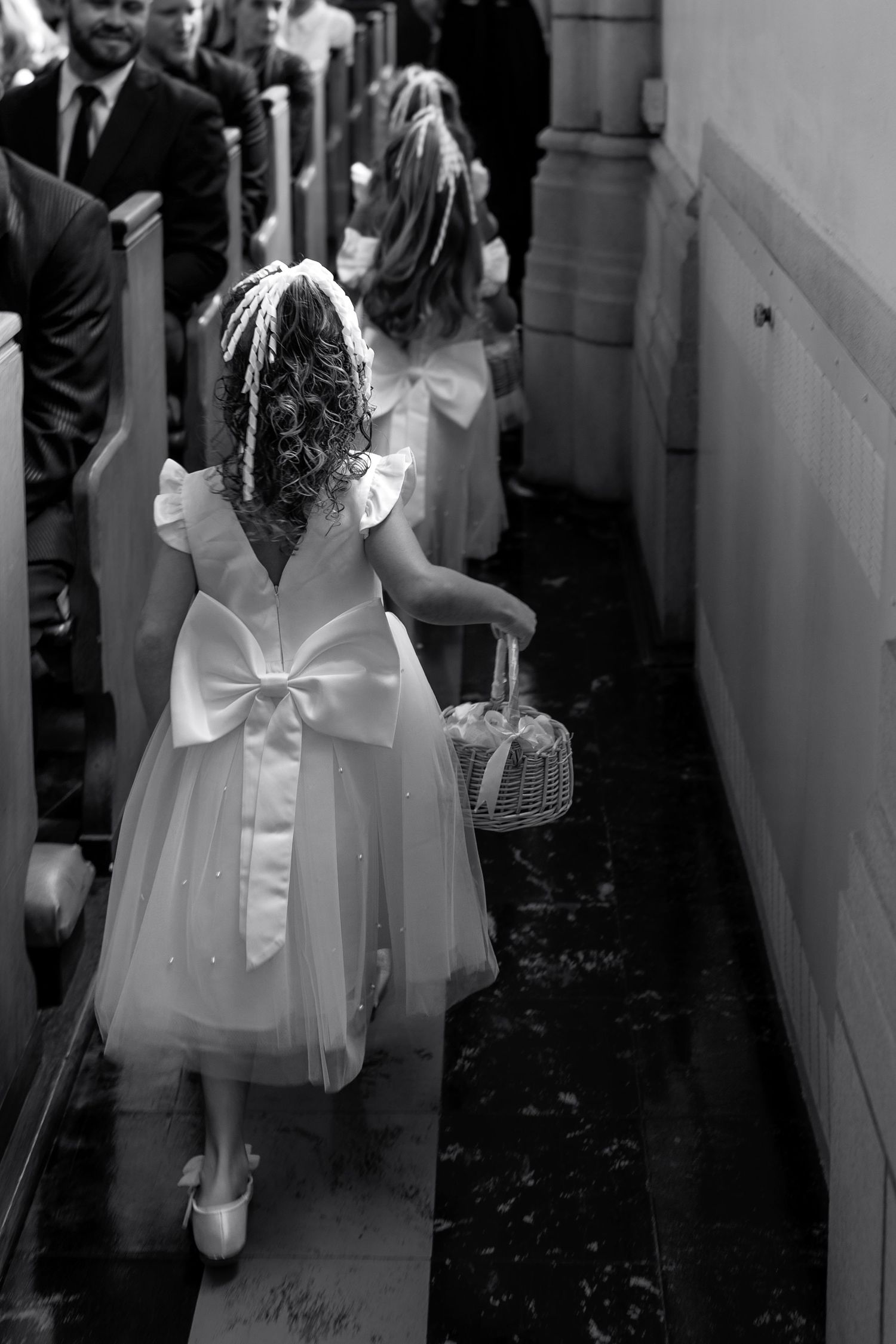 Young flower girl in white dress with bow walking down church aisle.