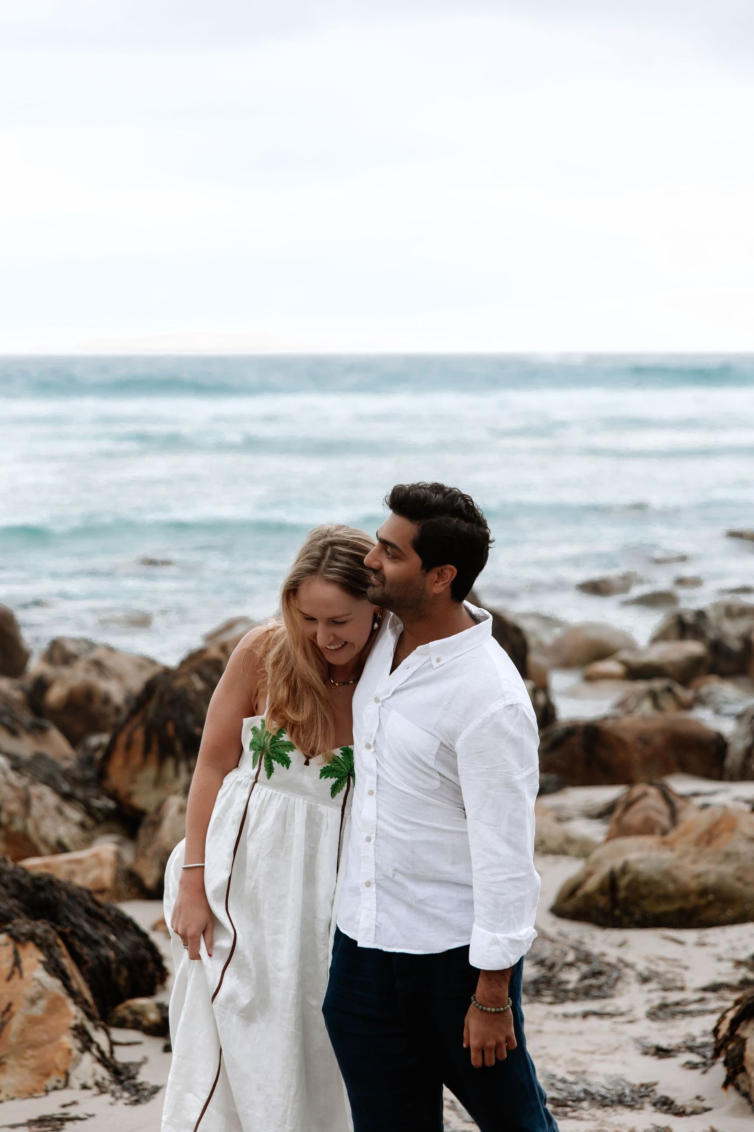 A couple shares a romantic moment on a rocky beach with crashing waves and ocean in the background.