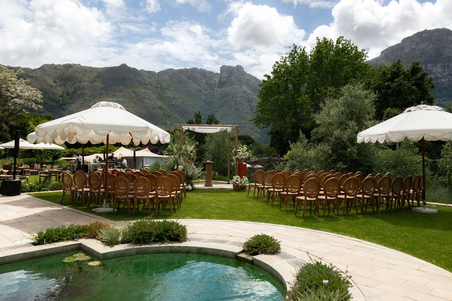 Panoramic view of an elegant outdoor wedding venue with mountain backdrop, pool, and white umbrellas set up for ceremony.