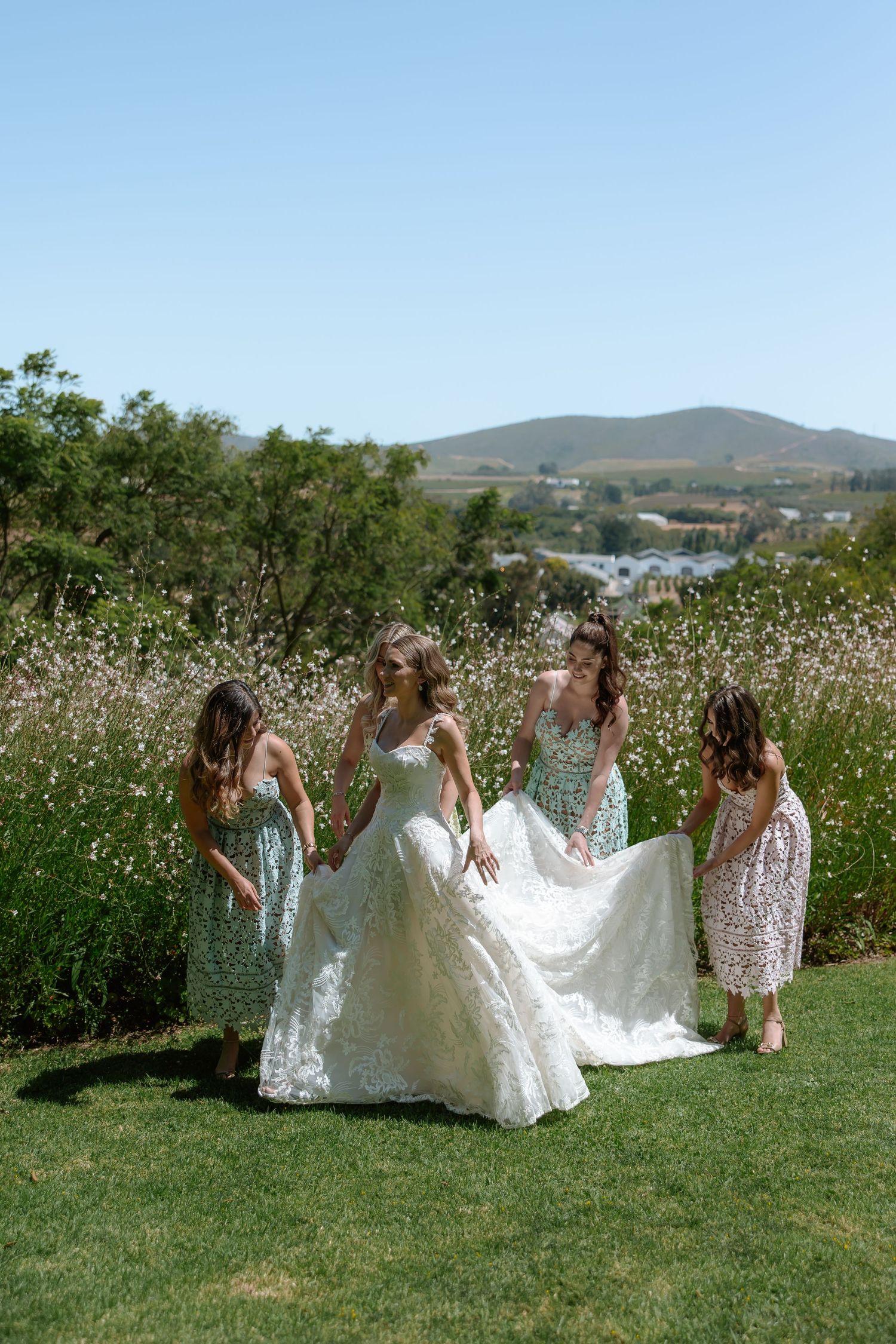 Group in floral dresses running through sunny meadow with wedding gown trailing behind.