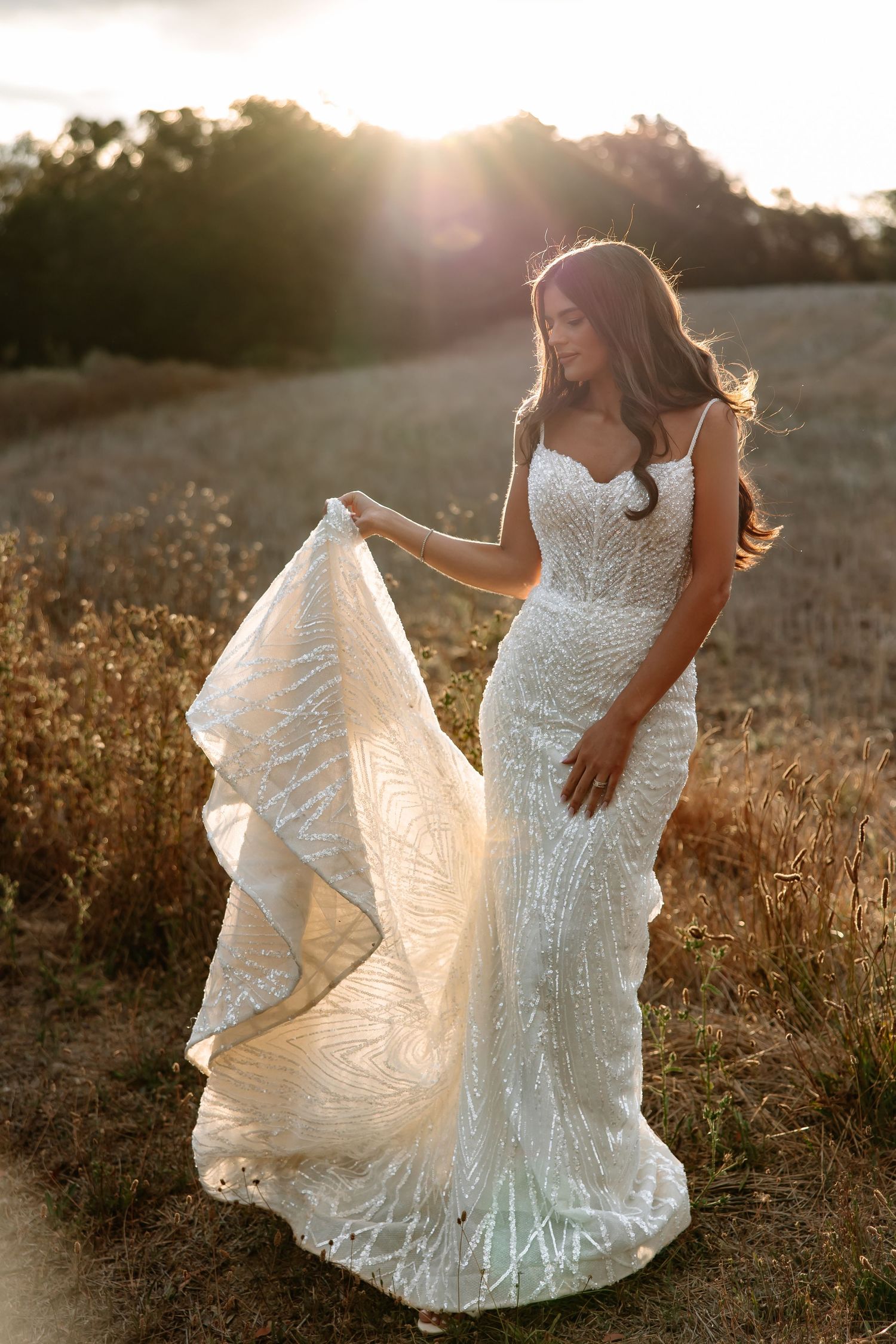 A stunning lace wedding dress catches golden sunlight in a field as its train flows elegantly in the breeze.