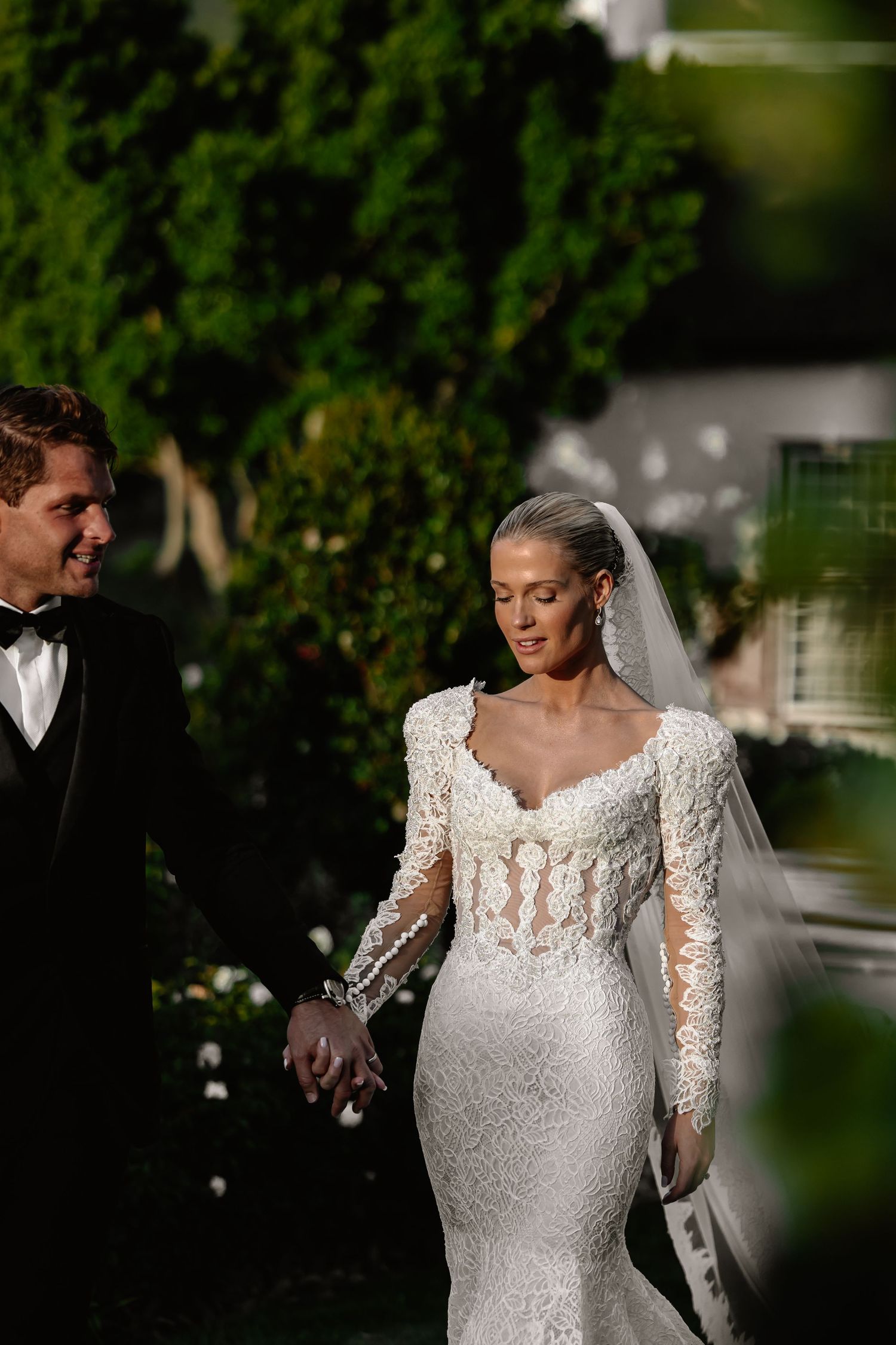 Romantic wedding couple walking together outdoors, bride wearing fitted lace gown with long sleeves.