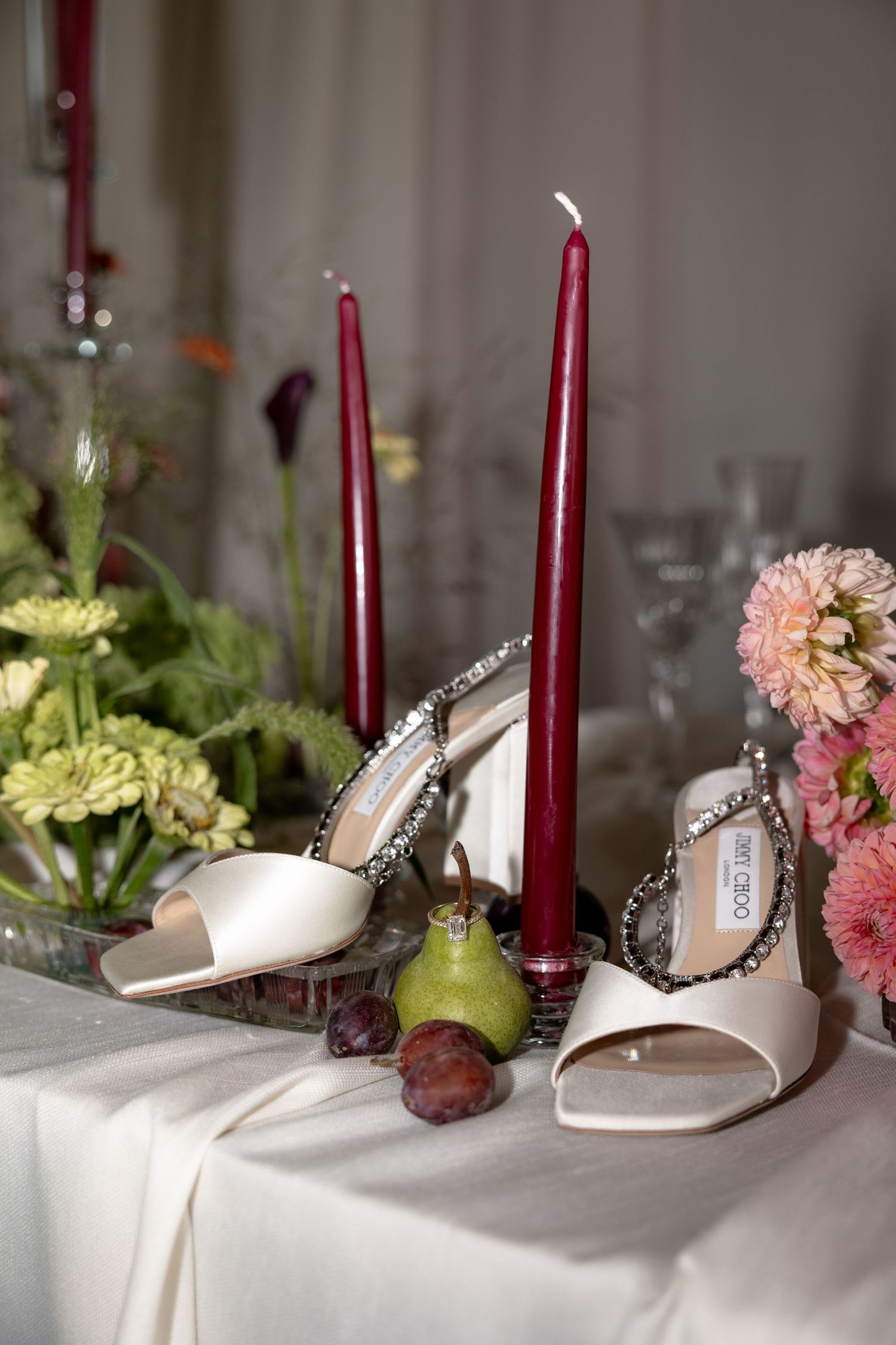 Elegant beige bridal shoes displayed on white tablecloth with burgundy candles and pink and green floral accents.