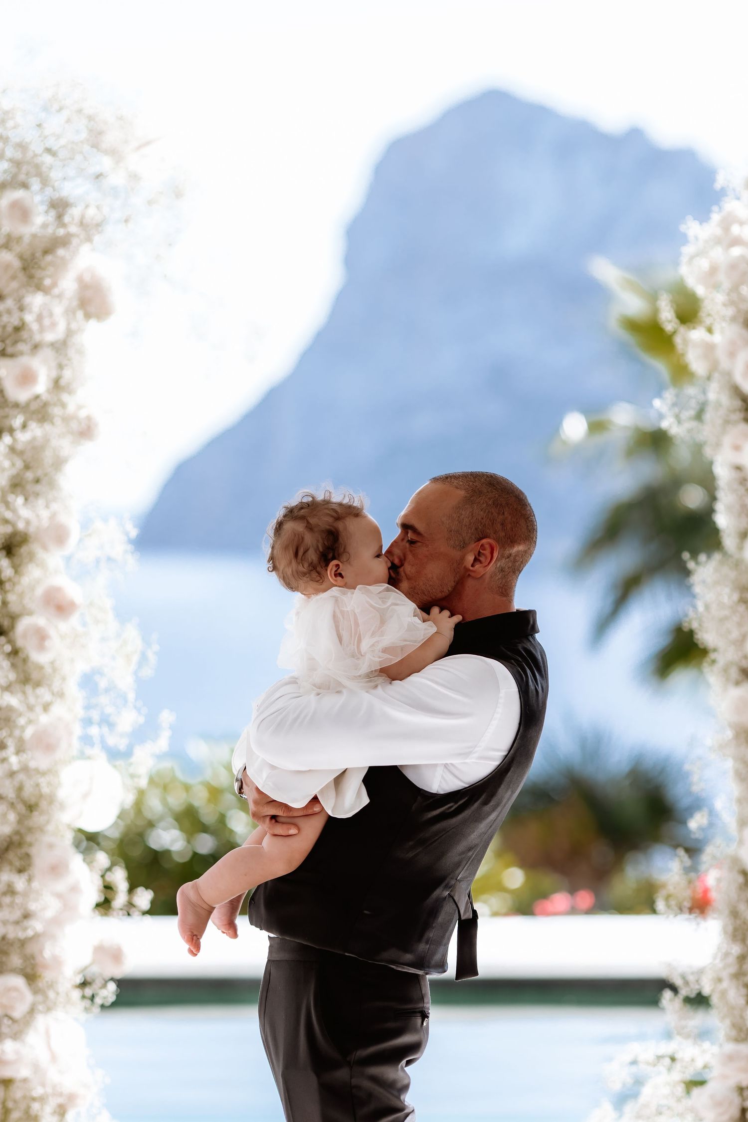 Romantic wedding couple embracing against a scenic ocean backdrop with mountain views and floral decor.