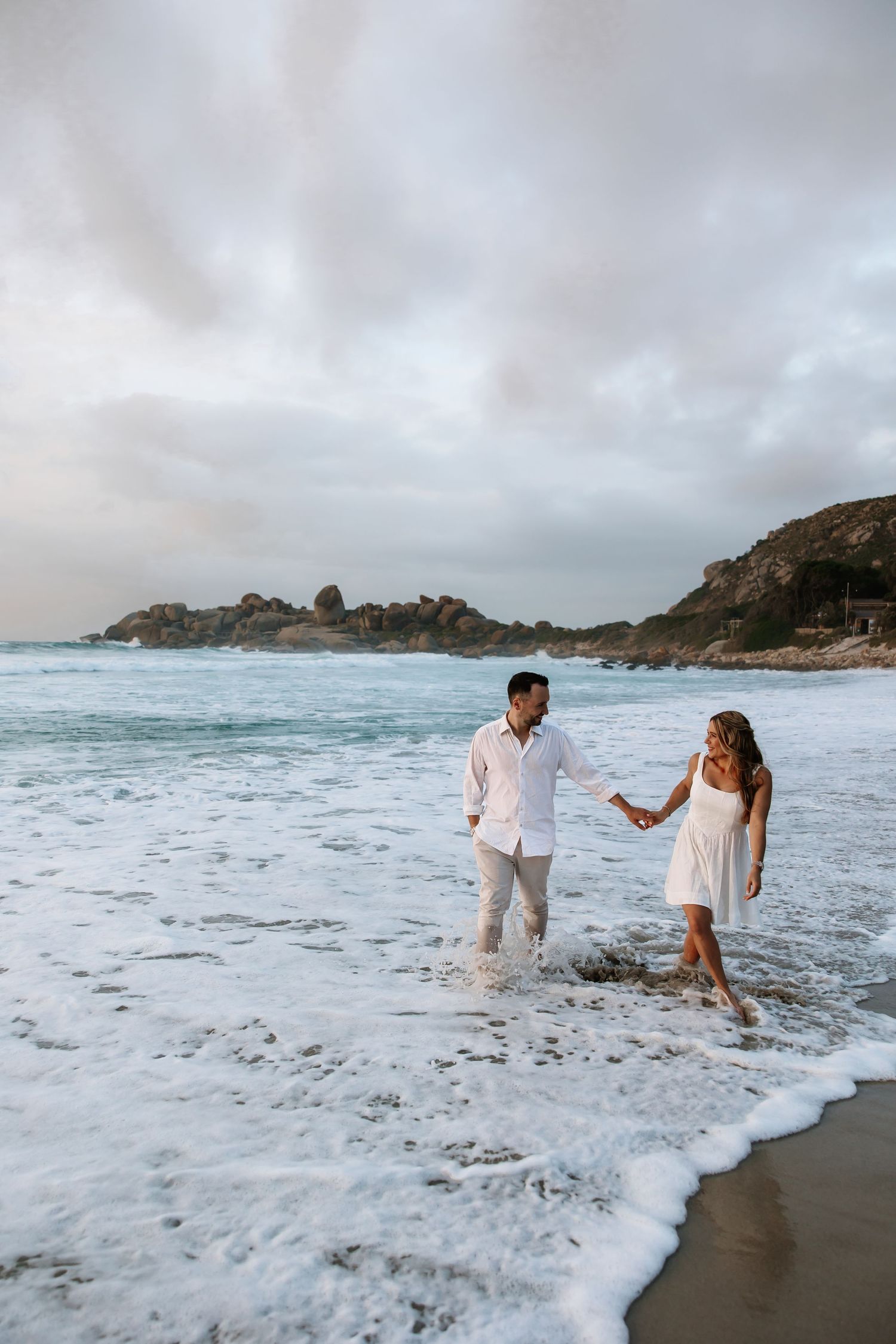 Couple in white outfits holding hands walking along a scenic beach with ocean waves and rocky cliffs.