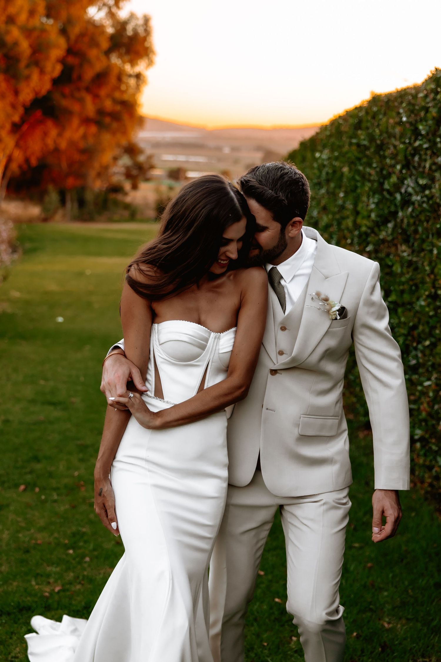 Romantic couple in white wedding attire embracing during golden hour sunset in a garden setting.