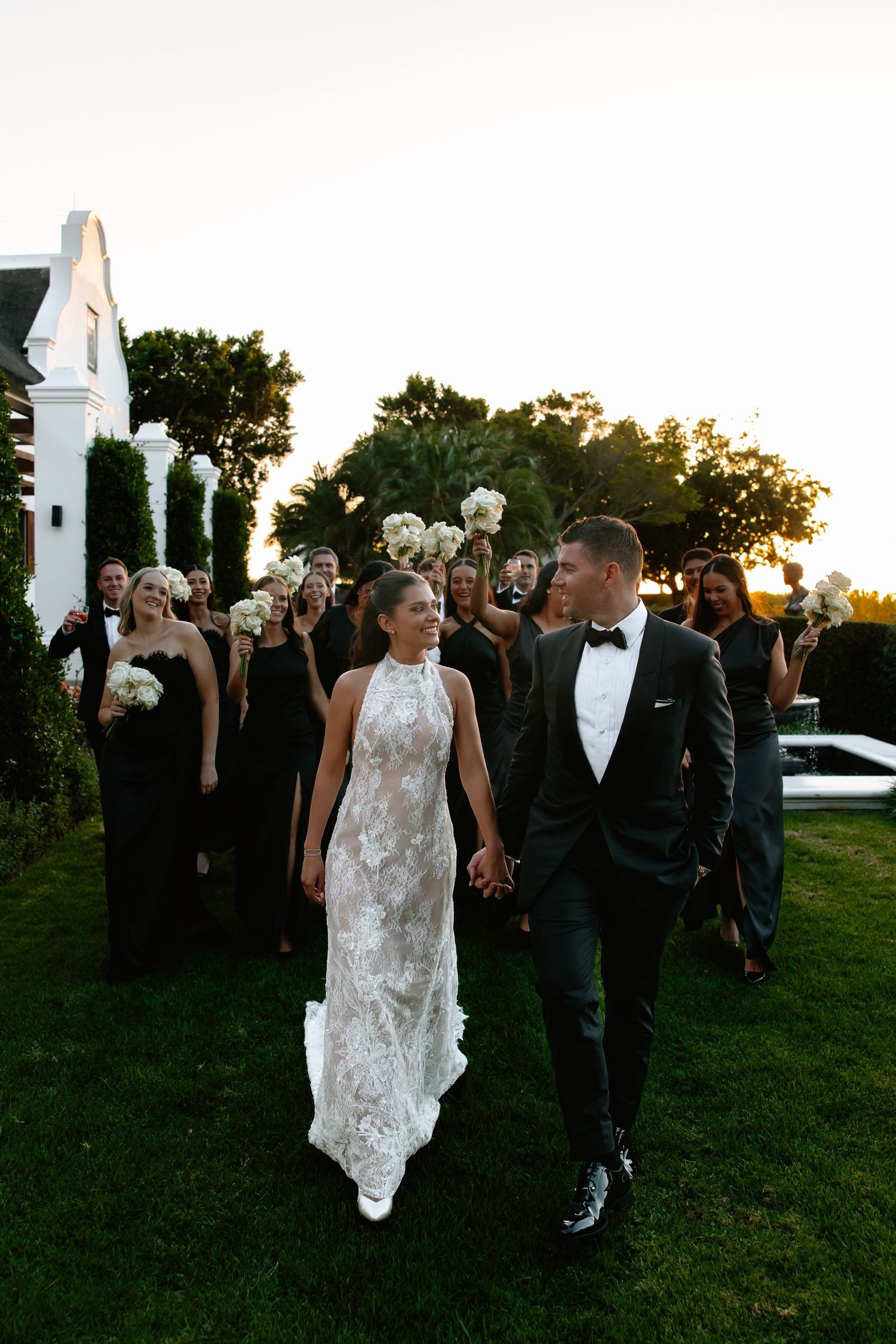 Newlywed couple walks hand in hand during golden hour at an outdoor wedding reception with guests in background.