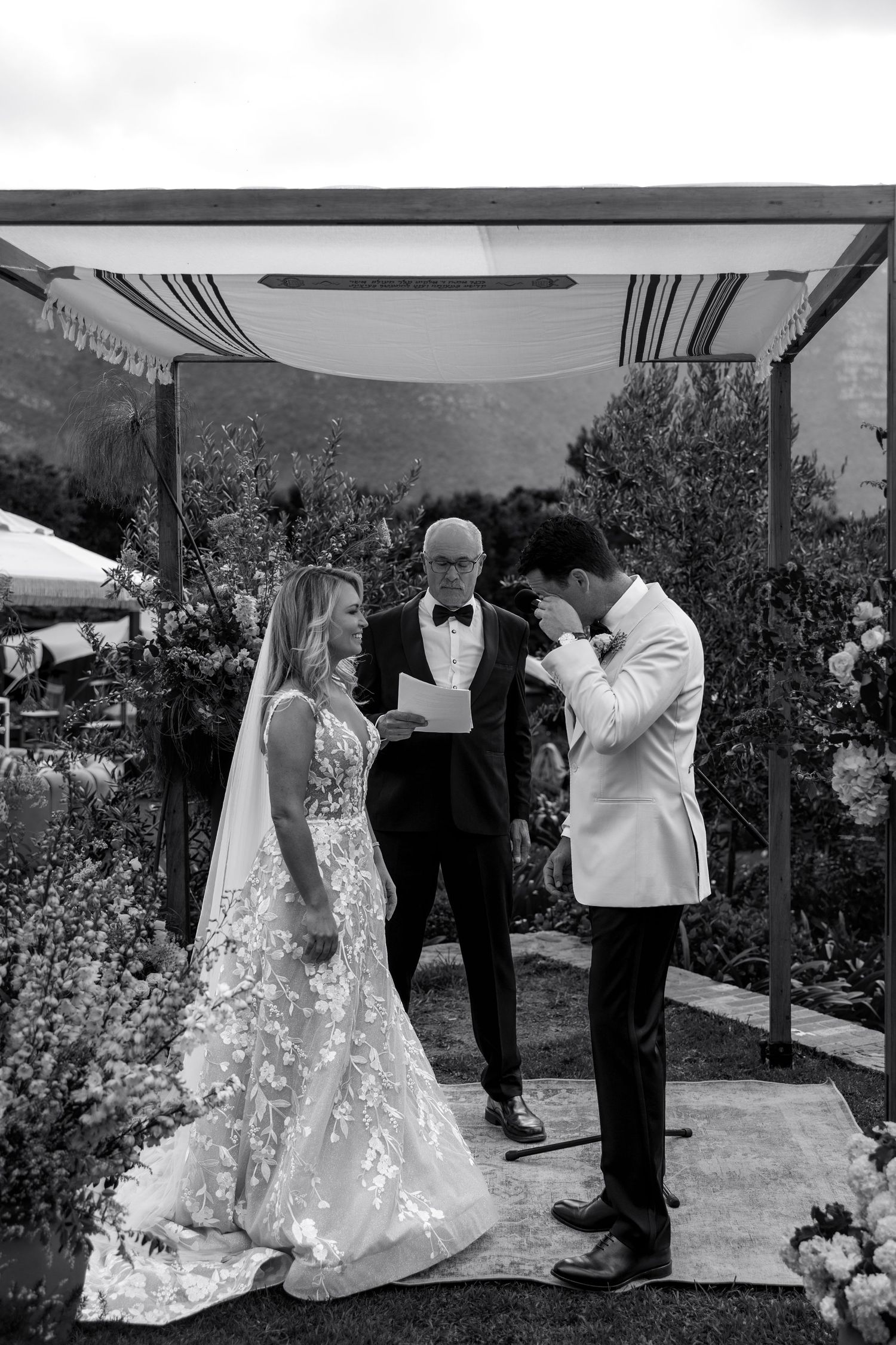 A black and white outdoor wedding ceremony takes place under a canopy with mountains visible in the background.