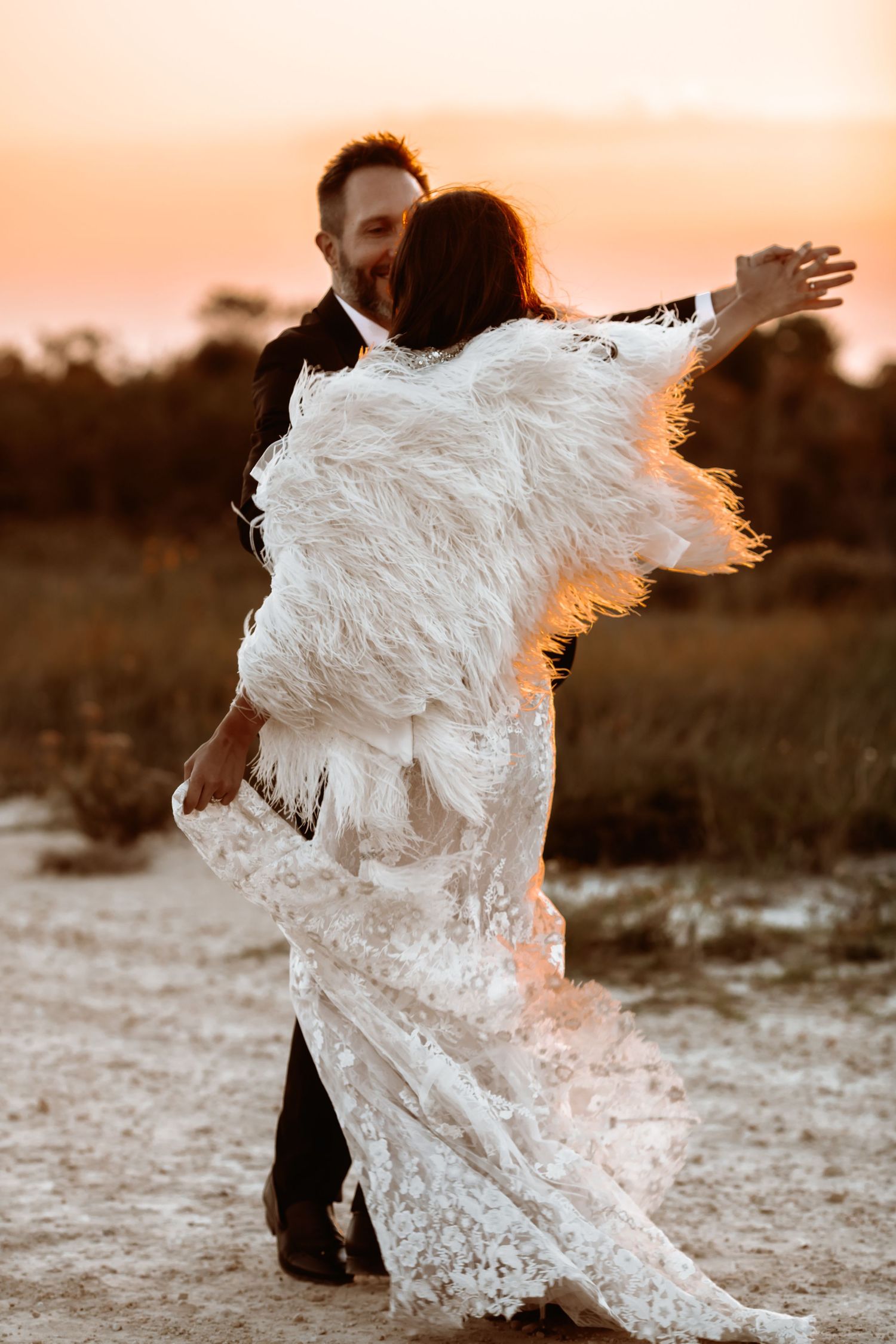 A flowing white lace wedding dress twirls dramatically against a sunset backdrop in an outdoor setting.