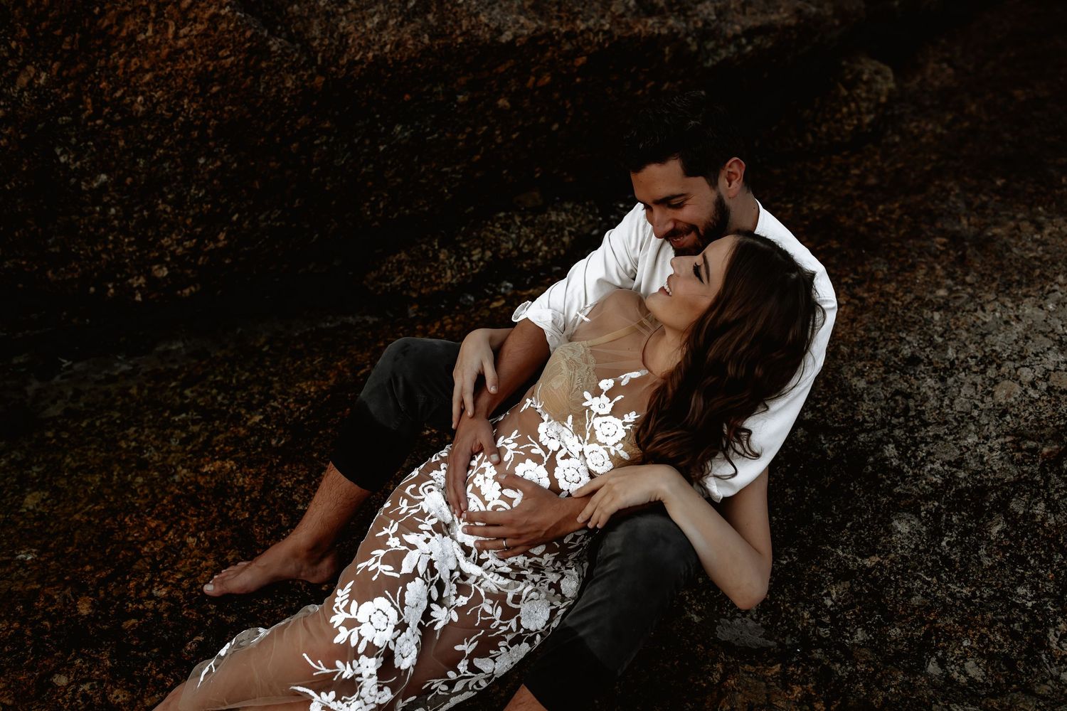 A couple reclines together on a white lounge chair against a dark textured background.