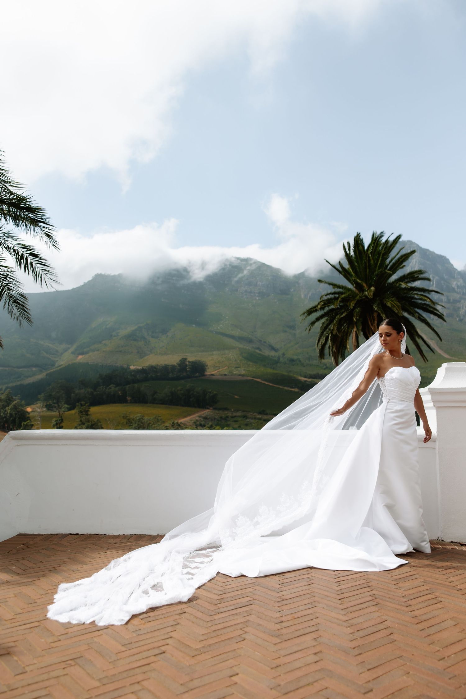 A flowing white wedding dress with a long train against a mountain landscape backdrop on a tiled terrace.