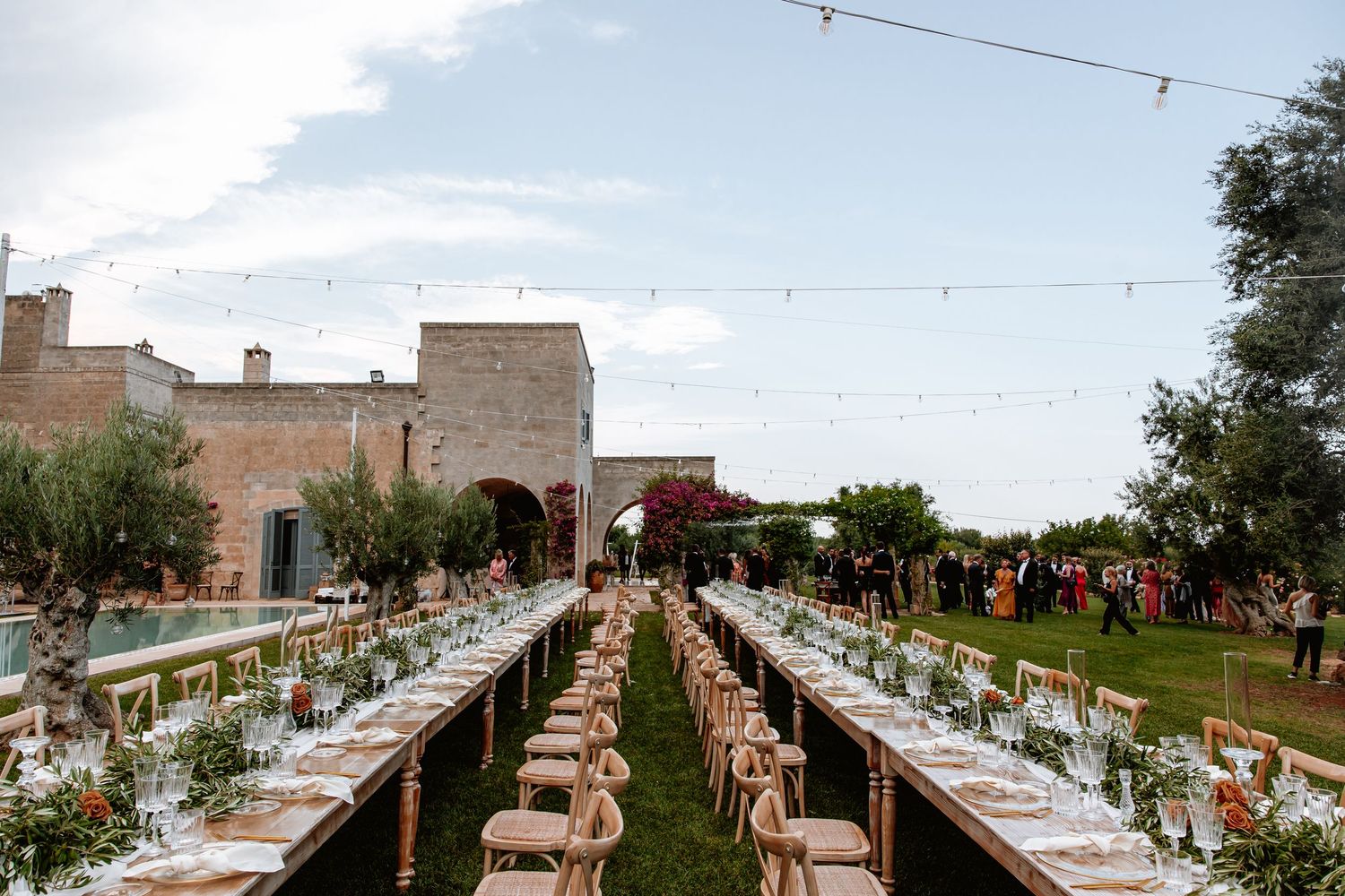 Long banquet tables set up outdoors with string lights and rustic decor for an elegant wedding reception.