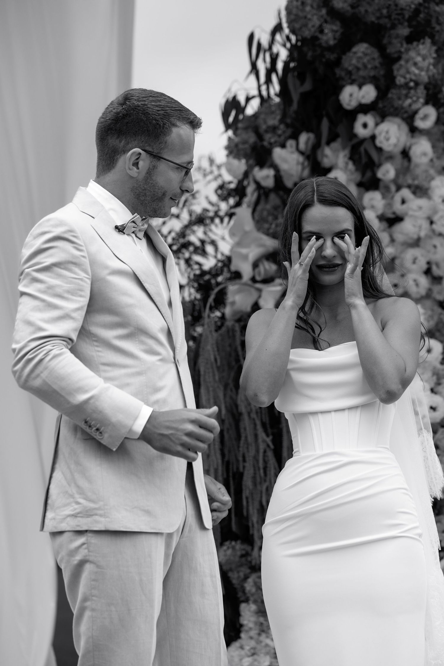 Black and white photo of a couple in white wedding attire sharing an emotional moment in front of floral decorations.