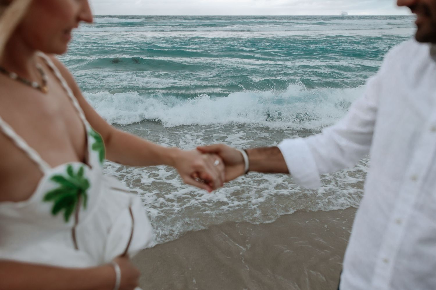 Couple walking along the beach holding hands in white wedding attire as ocean waves crash on the shore.