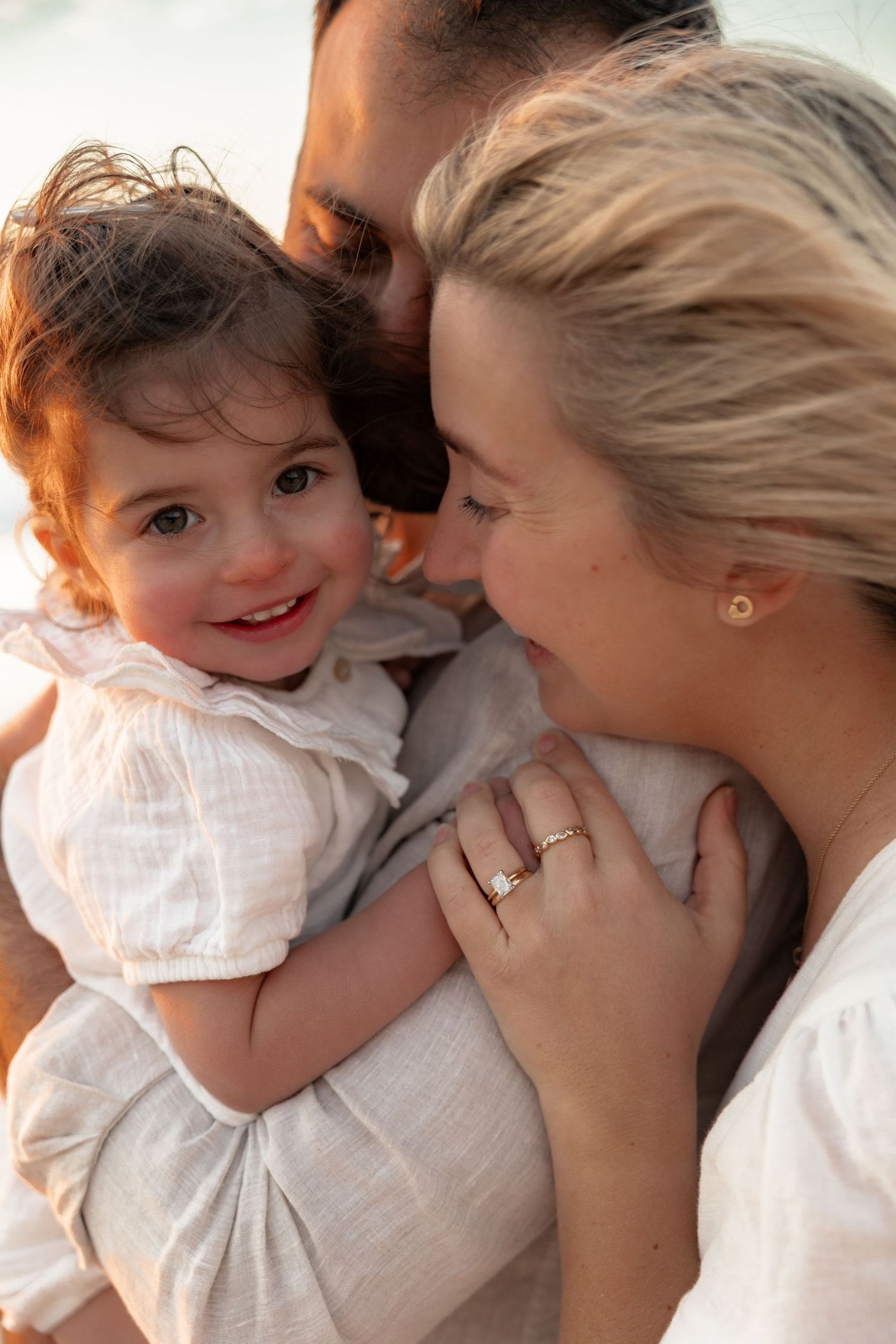 A couple lovingly holds their smiling toddler girl in white dress, sharing a tender family moment in warm light.