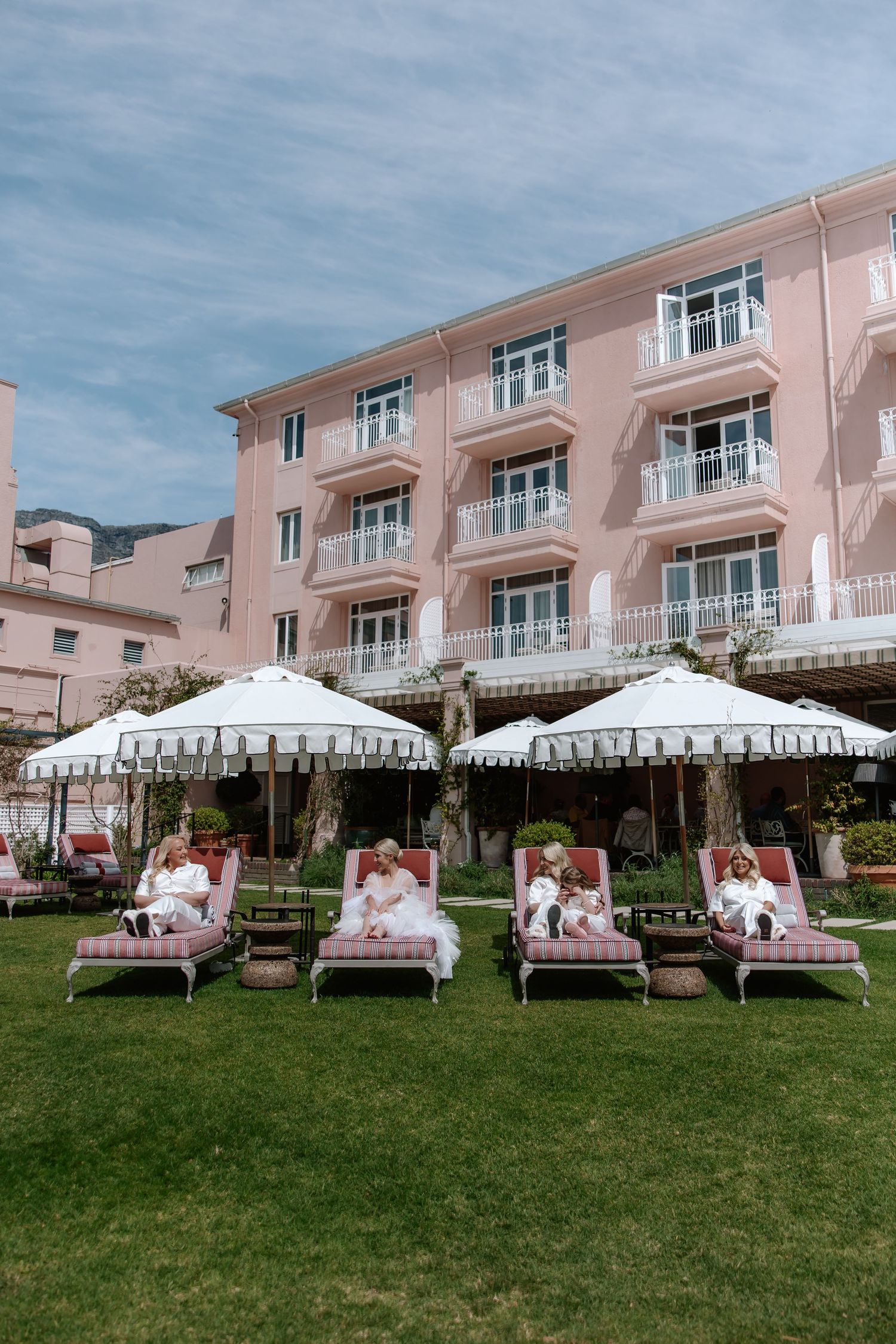 Pink beach chairs line the lawn outside a beachfront resort hotel under a blue sky.