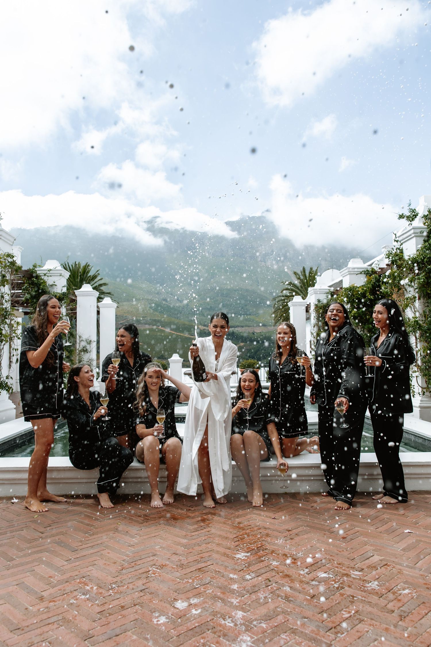 A wedding party stands on a terrace overlooking mountains while bubbles float through the air during a celebration.