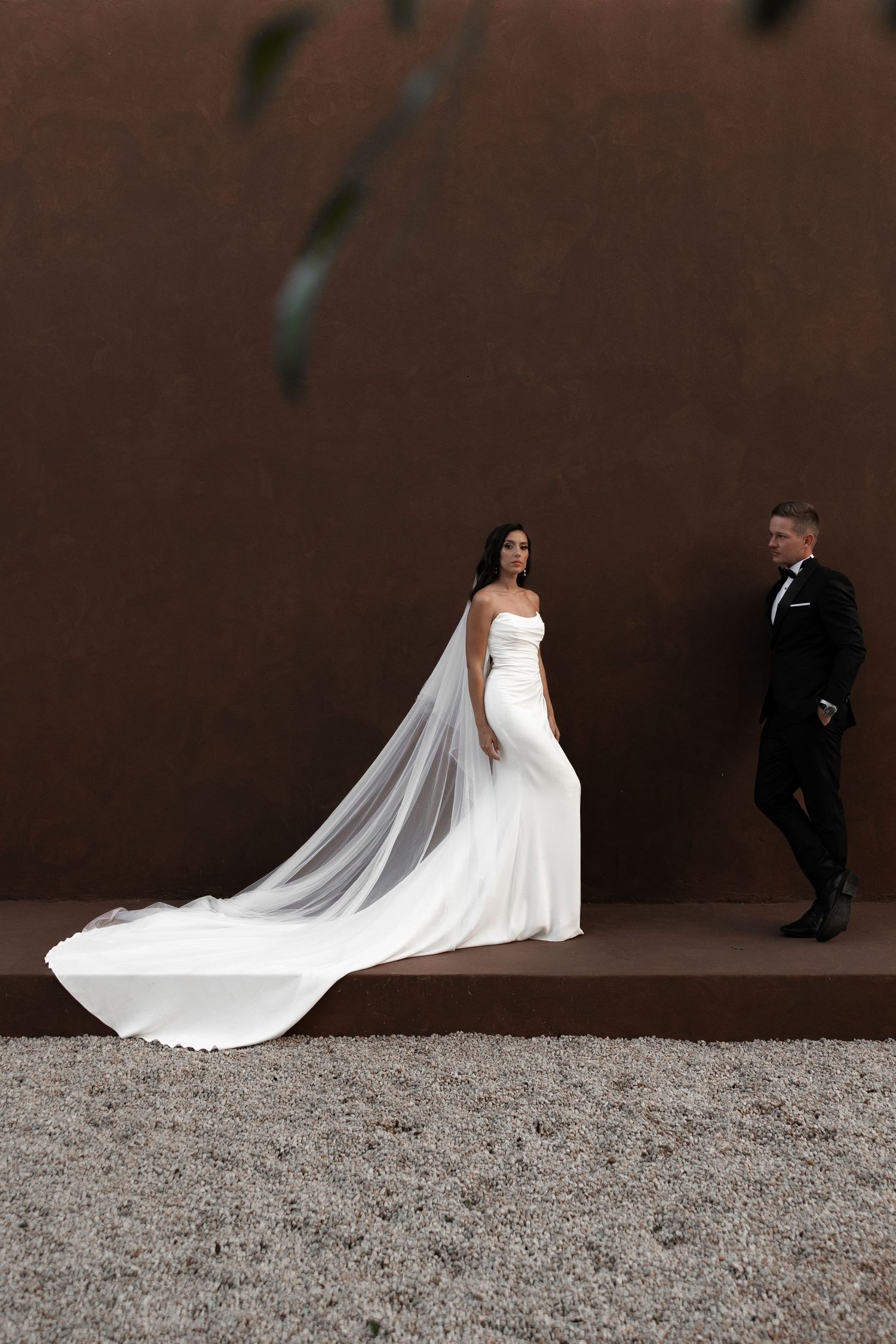 Wedding couple walking along a gravel path against a dark wall with flowing white veil.