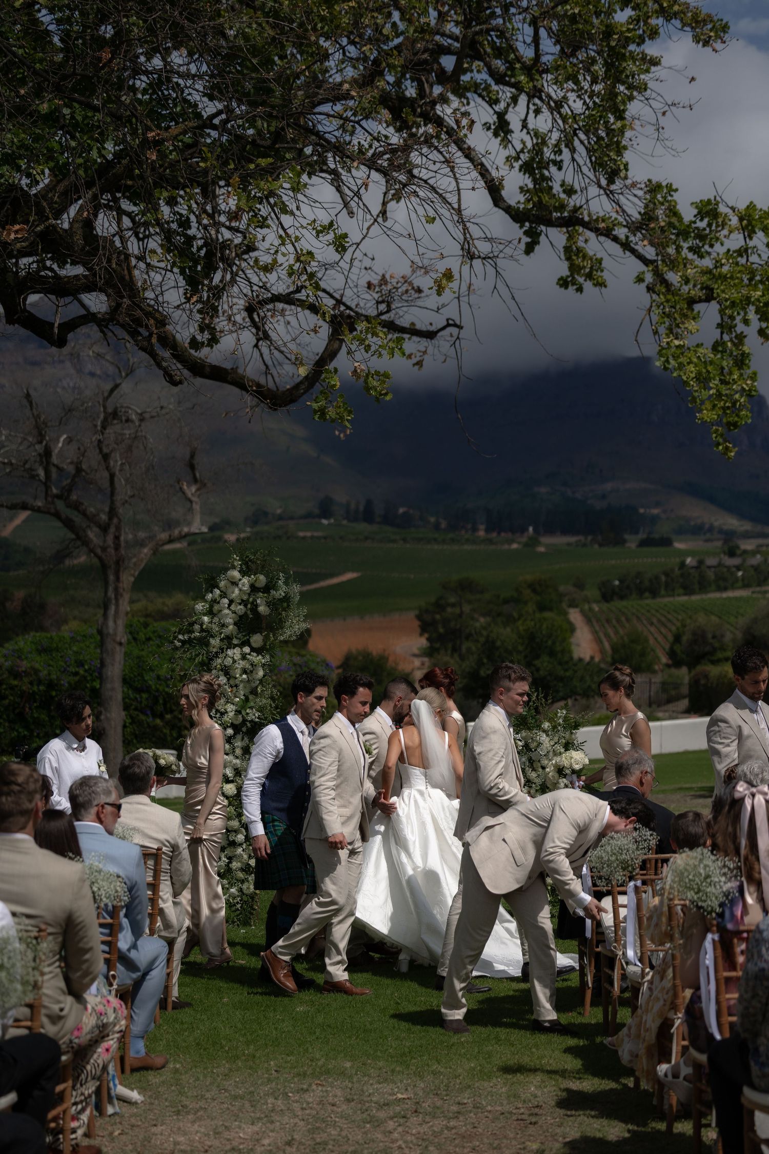 An outdoor wedding ceremony takes place on a hillside with mountains and rolling landscapes in the background.