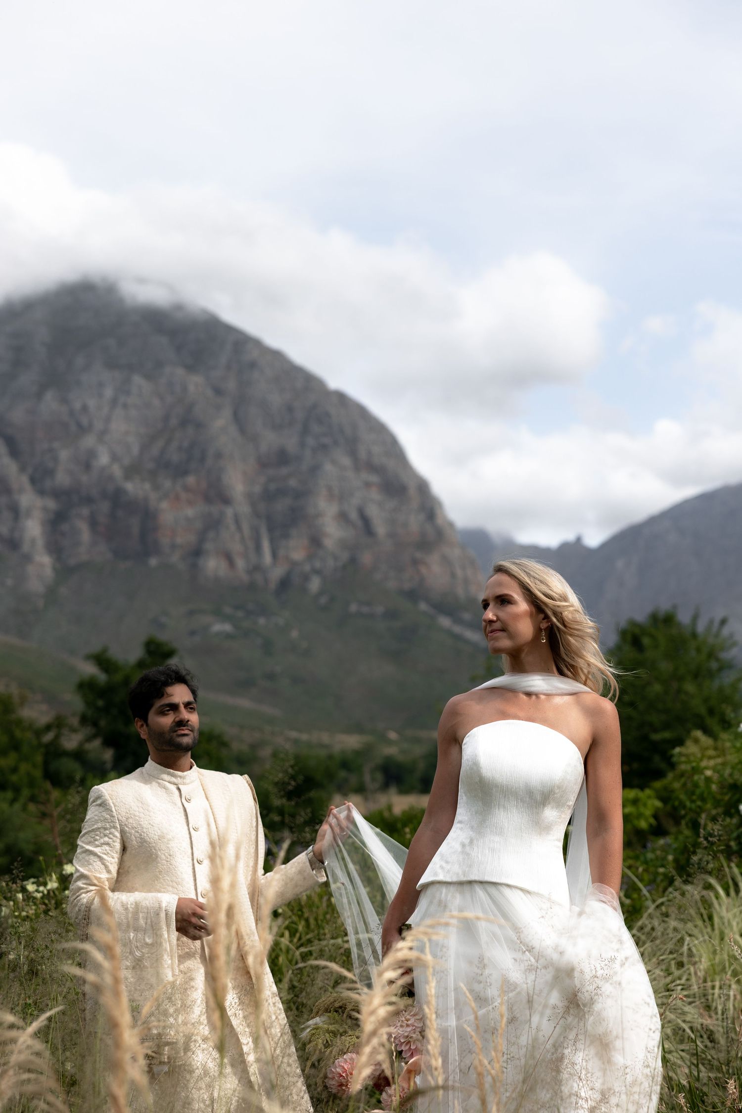 Bride in white gown and groom in ivory sherwani stand in a mountain field on their wedding day.
