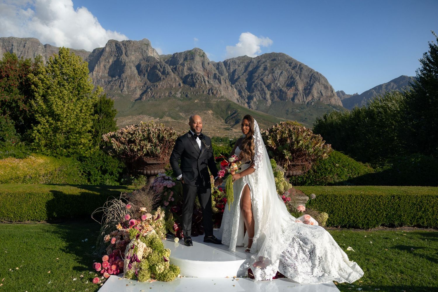 A scenic outdoor wedding ceremony in front of majestic mountain peaks with dramatic clouds and lush greenery.