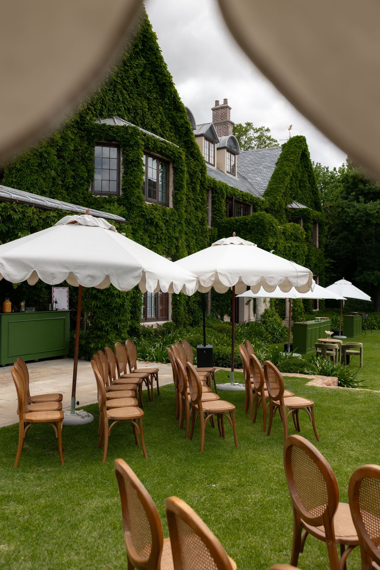 Outdoor garden seating with wooden chairs and white umbrellas arranged in front of an ivy-covered house with green shutters.