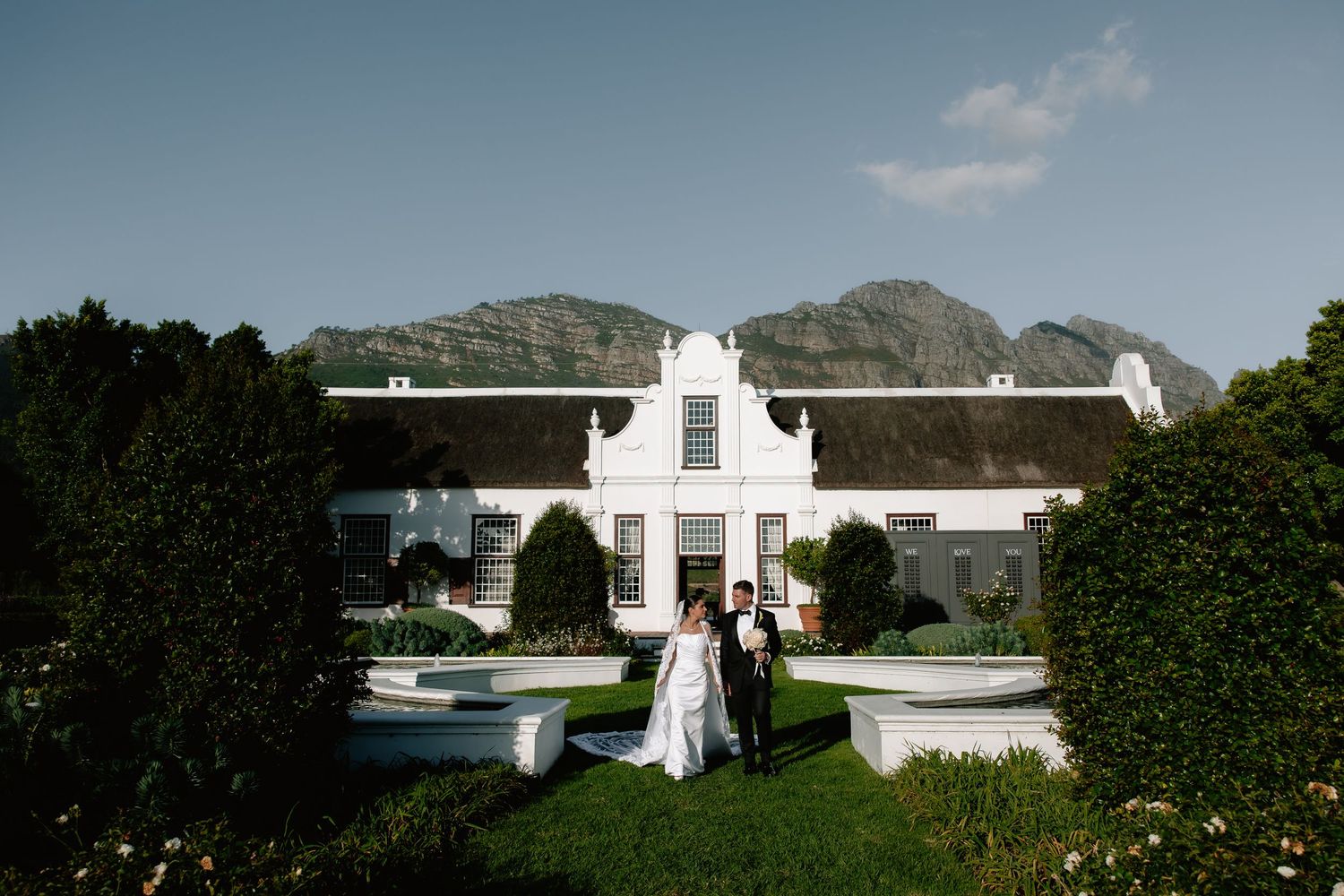 A couple stands in front of a grand Cape Dutch style mansion with white facade and mountains in the background.