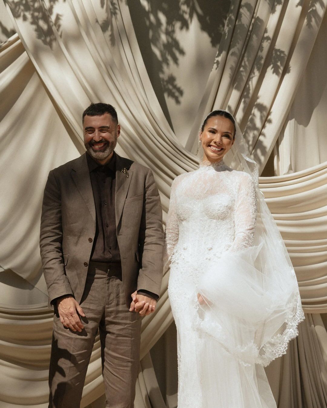 A couple poses together at their wedding, with the bride in a white lace gown and groom in a tan suit against draped fabric.