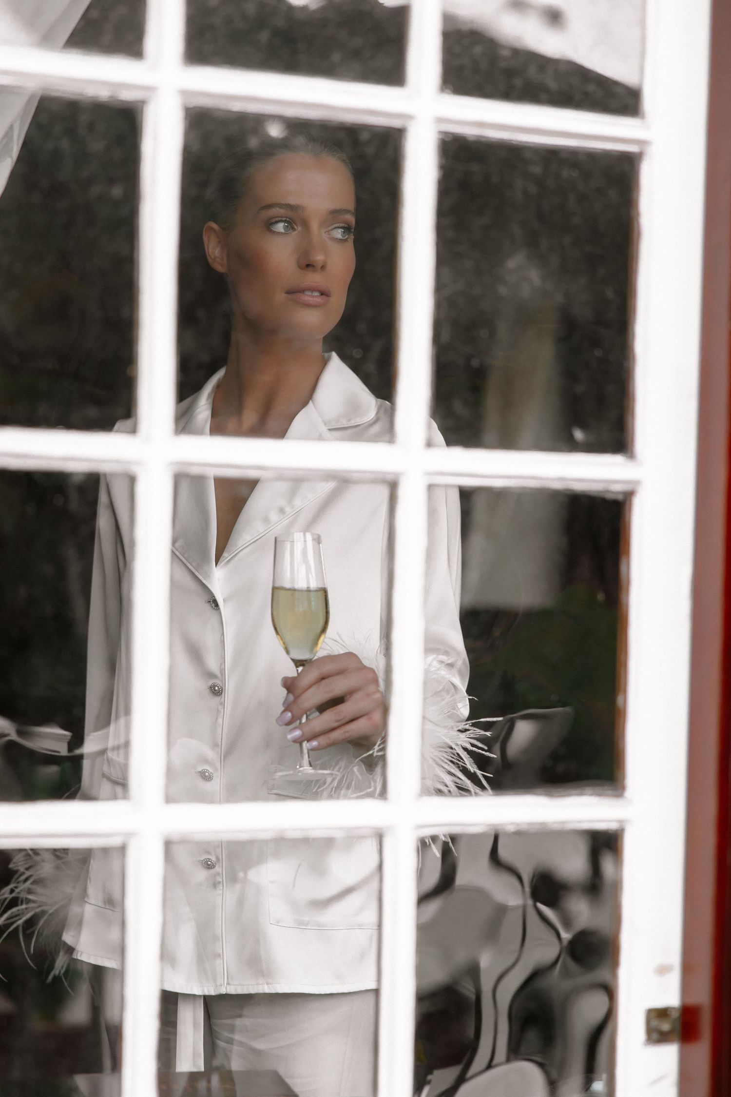 A white-clad figure is seen through window panes holding a champagne glass.