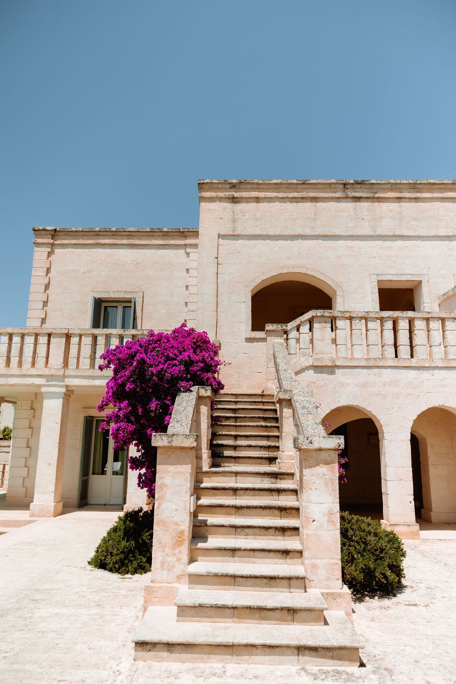 White stone staircase with vibrant purple bougainvillea flowers against a Mediterranean building facade.
