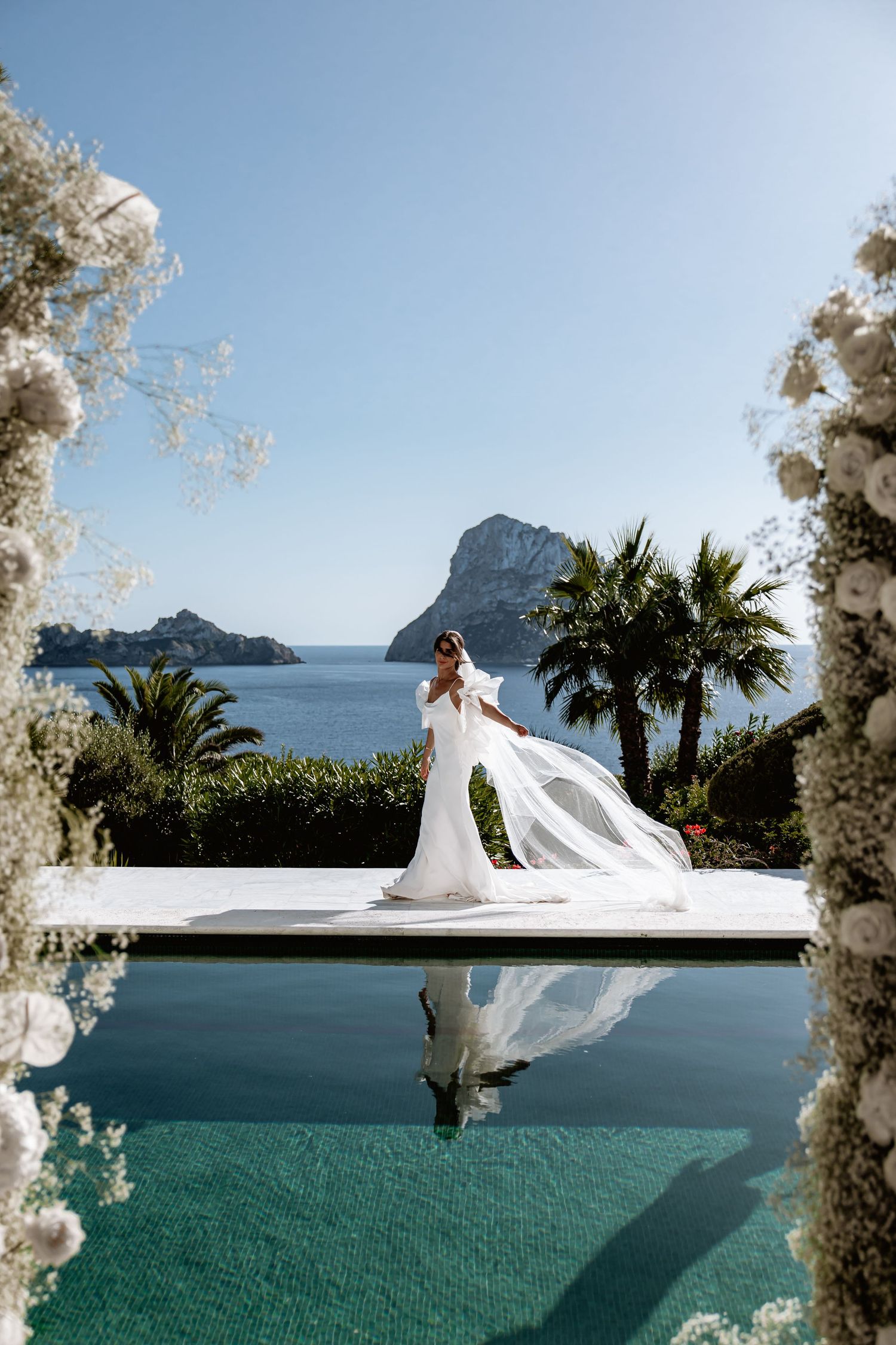 A bride in a flowing white dress stands on a platform overlooking the Mediterranean Sea and rocky coastline in Ibiza.
