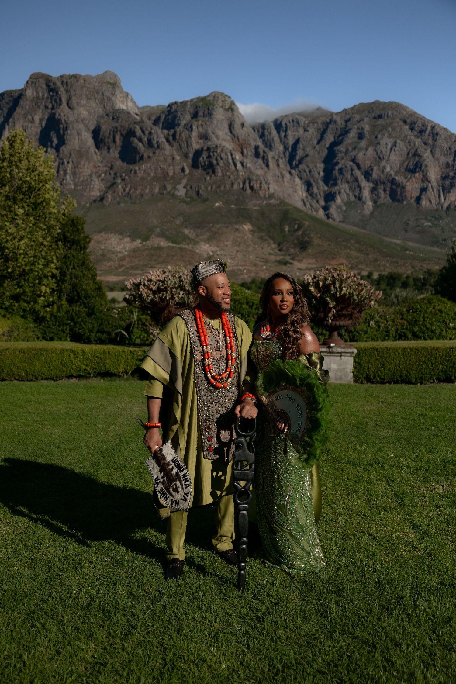 Two figures stand together on a grassy field with dramatic mountain peaks rising in the background.
