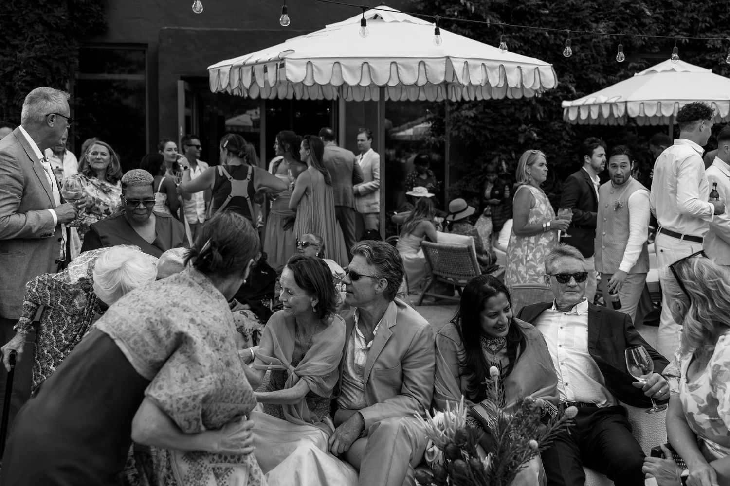 Black and white photo of elegantly dressed guests socializing outdoors under striped umbrellas at a garden party.