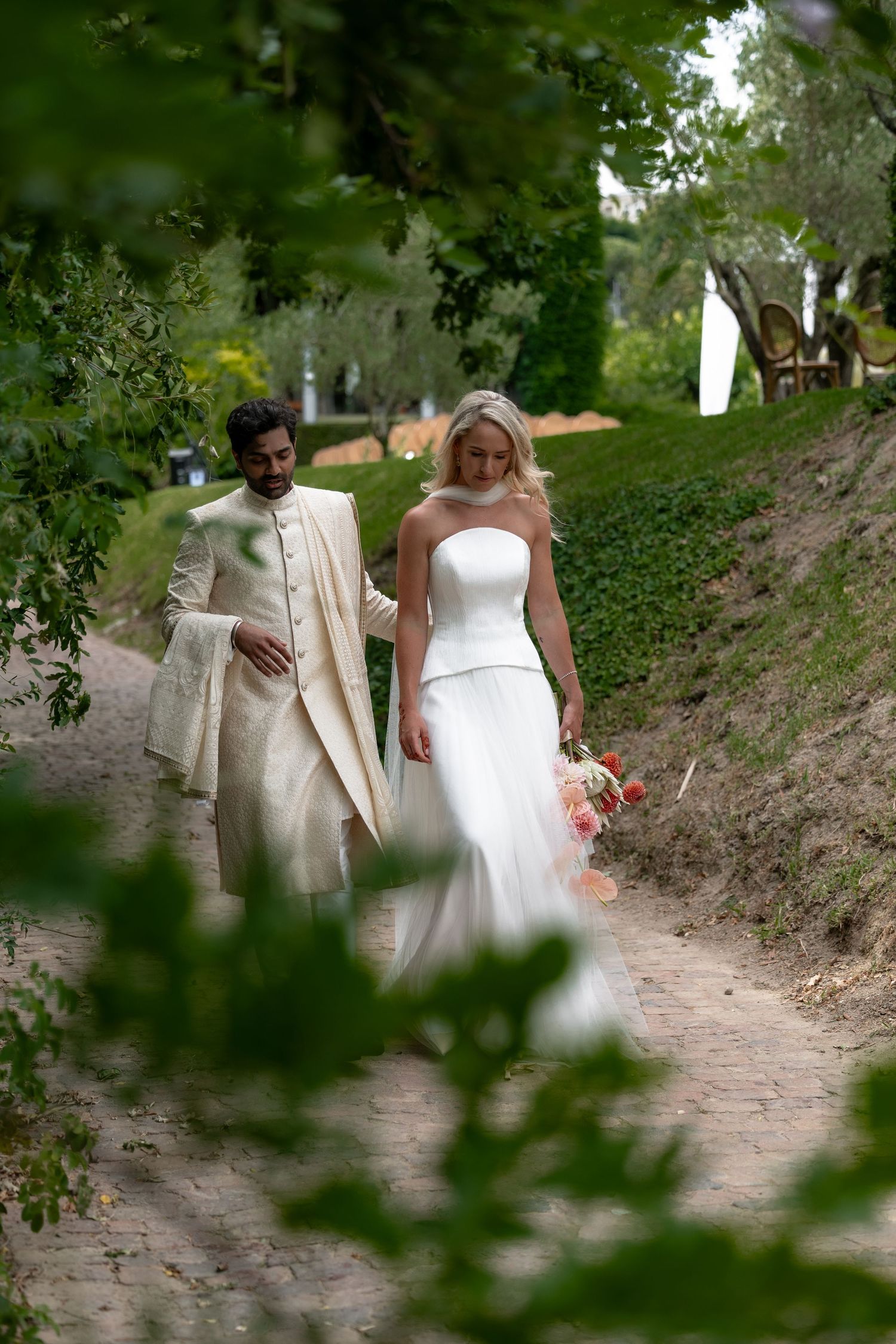 A couple walking down a scenic outdoor path in elegant white wedding attire.
