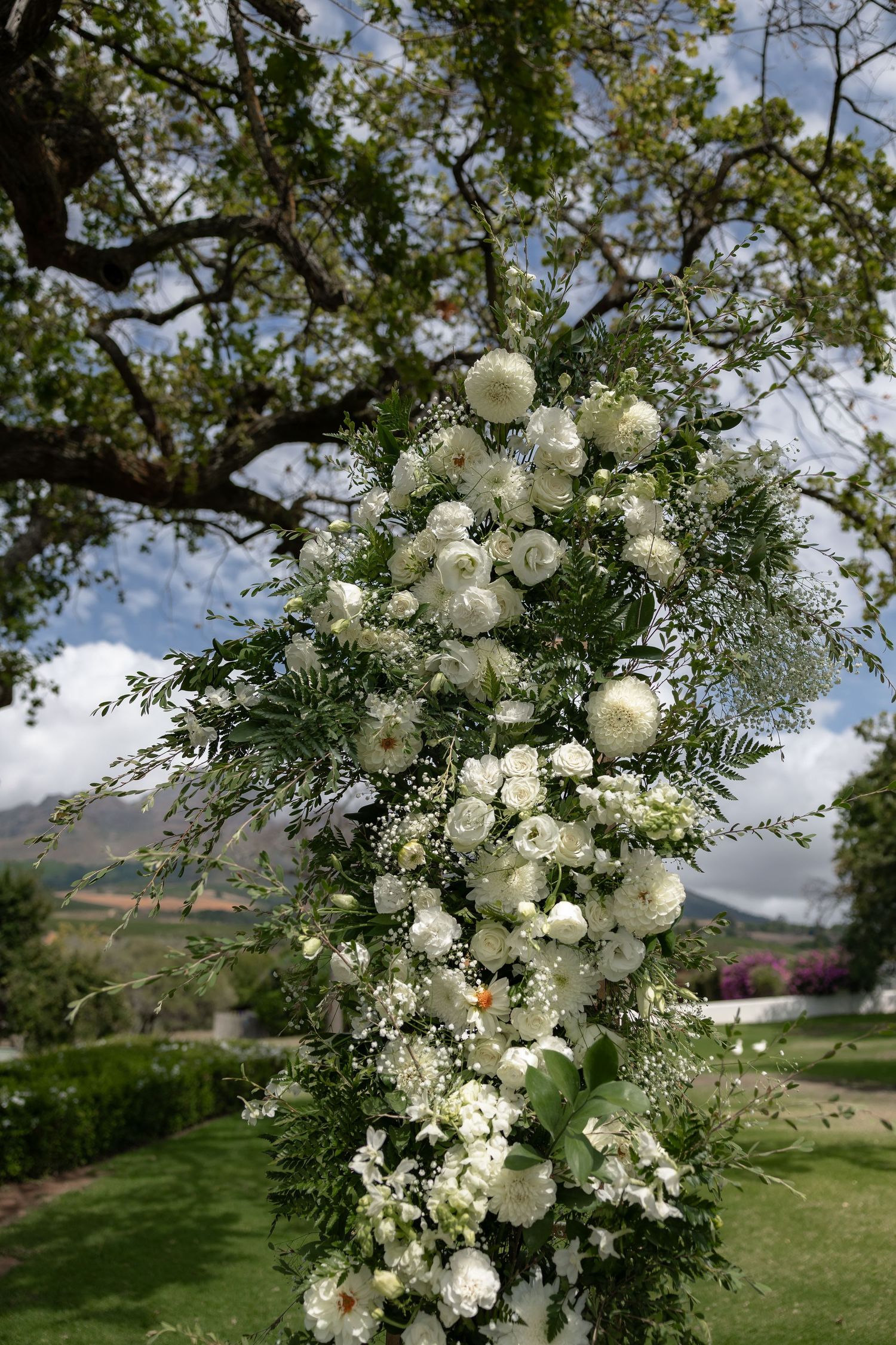 An elegant white floral arrangement with roses and greenery decorates an outdoor garden setting against a blue sky.