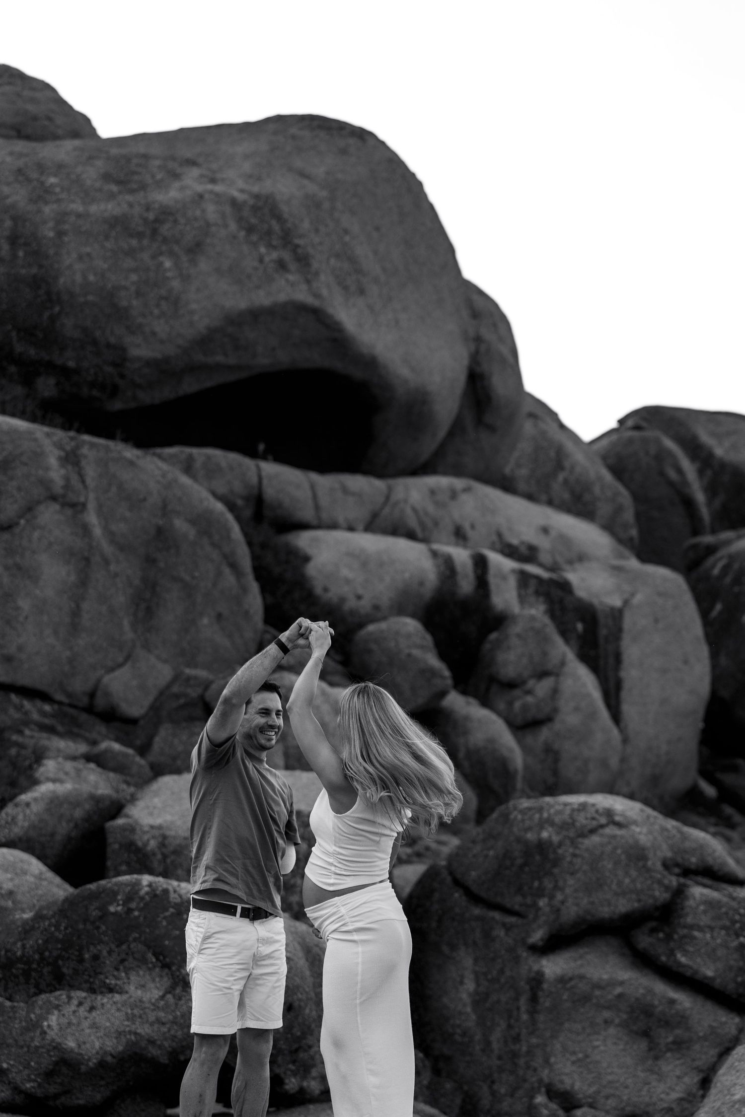 Black and white photo of a couple dancing joyfully among large granite boulders outdoors.