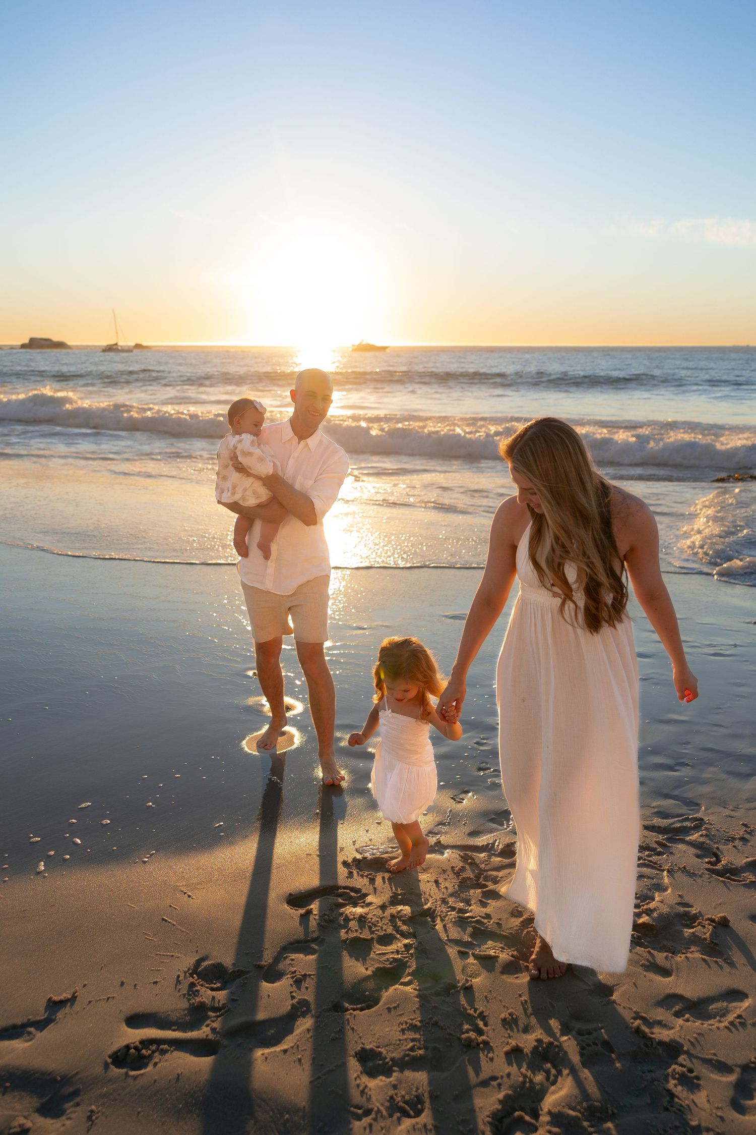 Family in white outfits walking on a sandy beach at sunset, parents holding children near ocean waves.