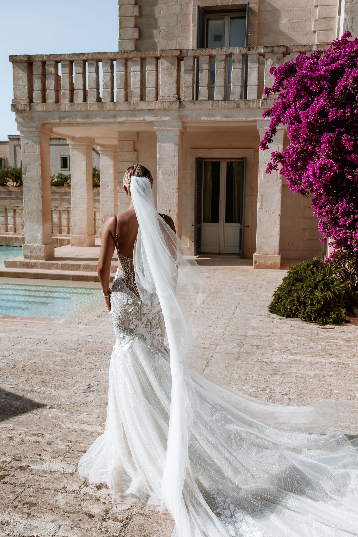 Bride in lace wedding gown with cathedral veil twirling on stone terrace near bougainvillea.