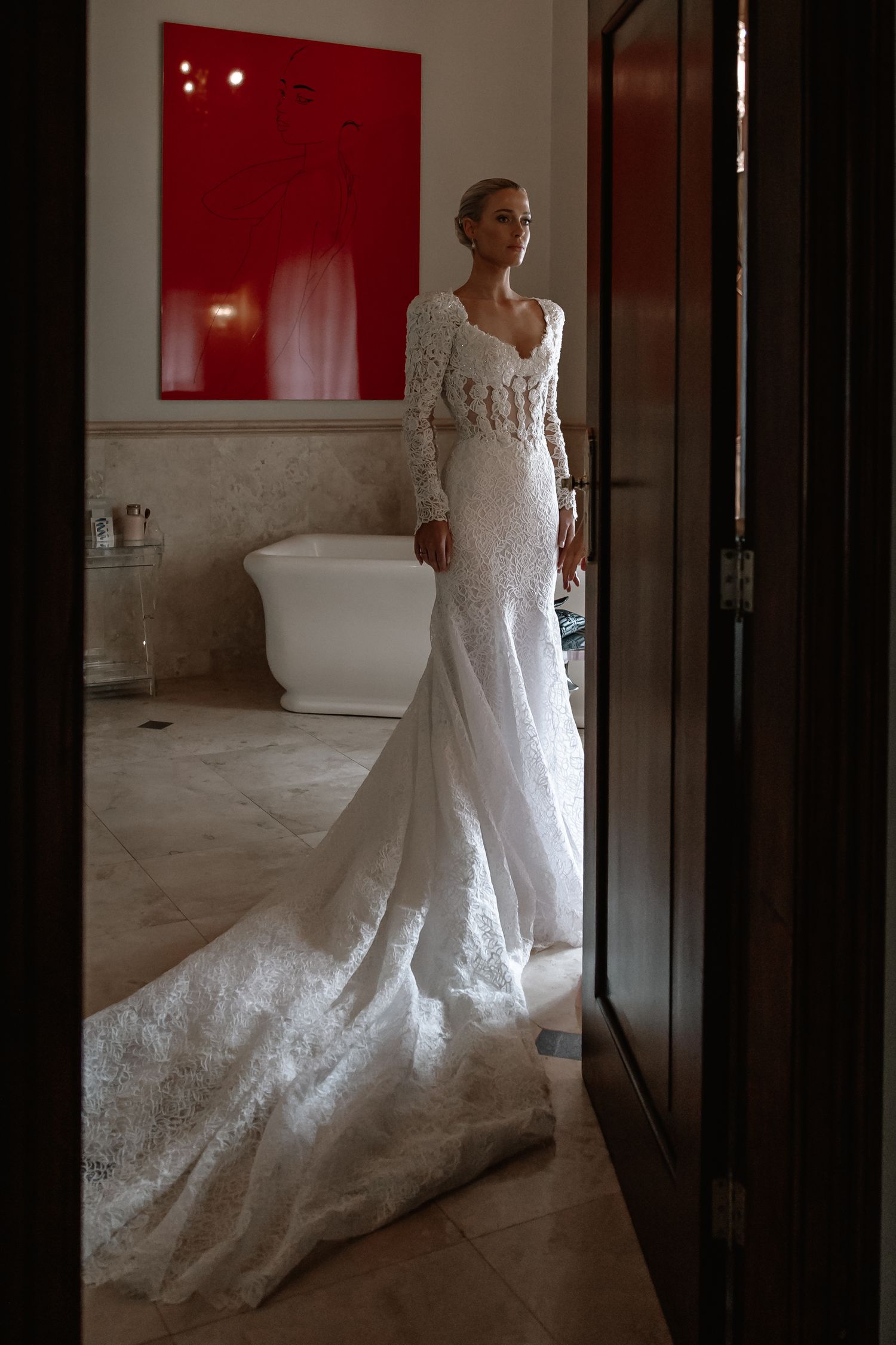 A white lace wedding gown with a dramatic train is photographed in a minimalist interior setting.