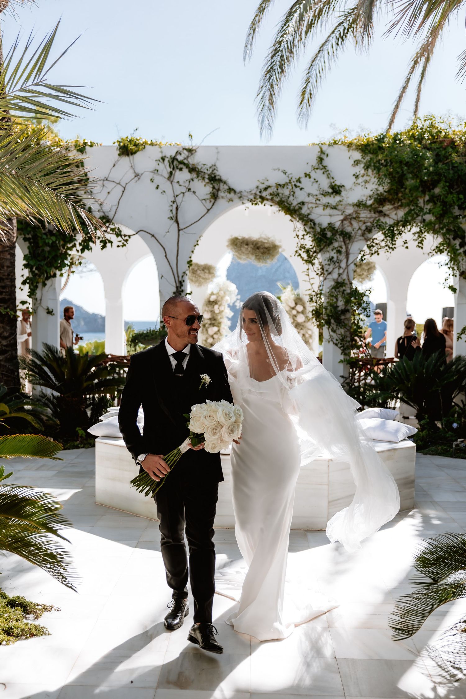 A wedding ceremony takes place in a Mediterranean-style venue with white arches and palm trees overlooking the ocean.