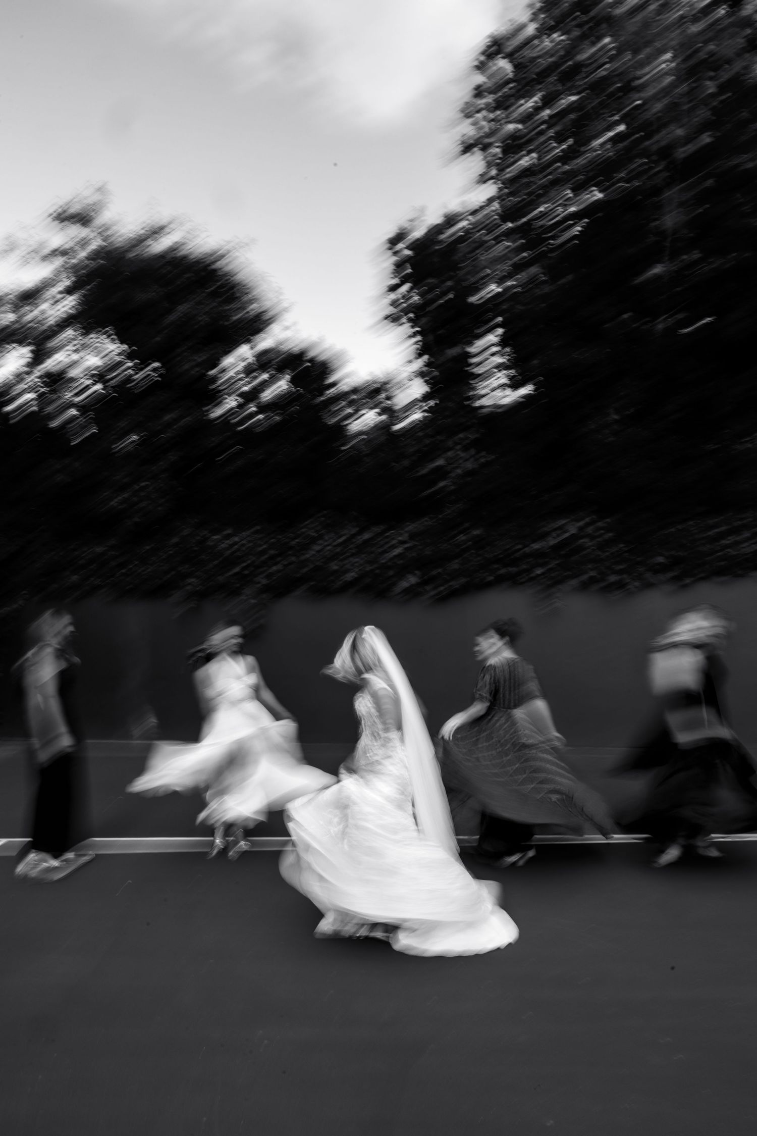 A black and white motion-blurred photograph shows people running and dancing in flowing dresses against dark trees.