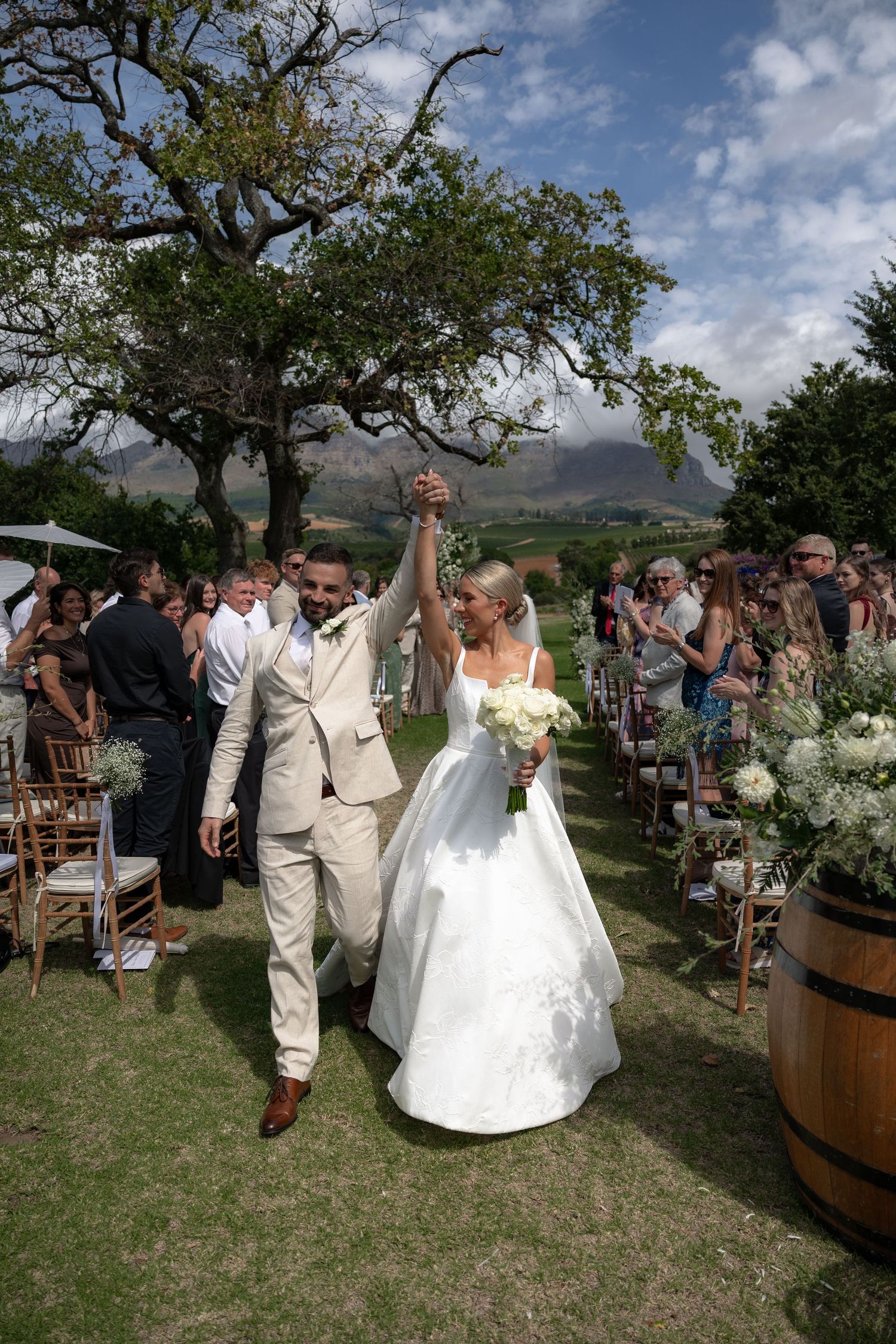 Outdoor wedding ceremony takes place on grassy hillside with scenic mountain views and rustic wine barrels as decor.