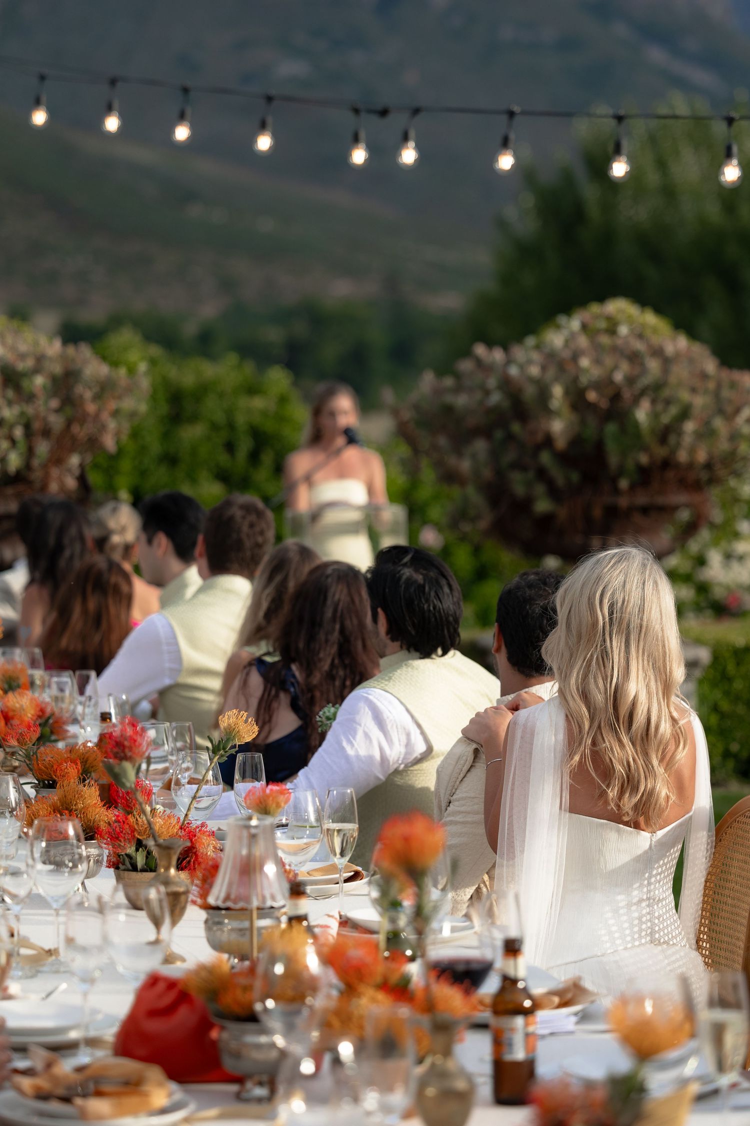 Outdoor wedding reception table set with string lights and orange floral centerpieces.